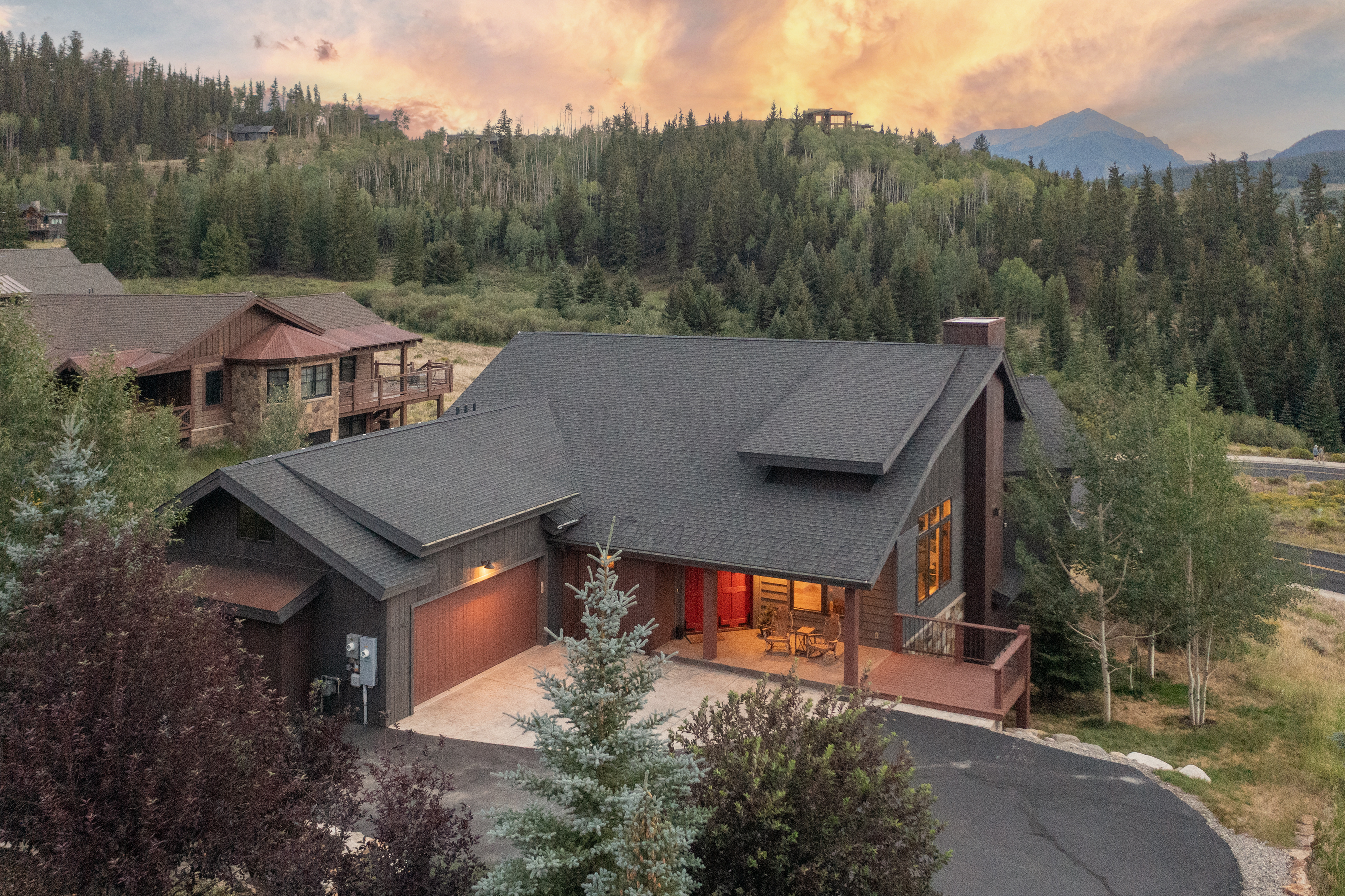 This aerial perspective captures a mountain-style home at dusk, set against a dramatic, colorful sunset sky. The residence features dark wood siding, a multi-gabled roof, and a prominent stone-clad chimney breast, with a covered front porch and an attached garage. The surrounding landscape is filled with dense evergreen trees and aspen groves, creating a serene and secluded atmosphere.