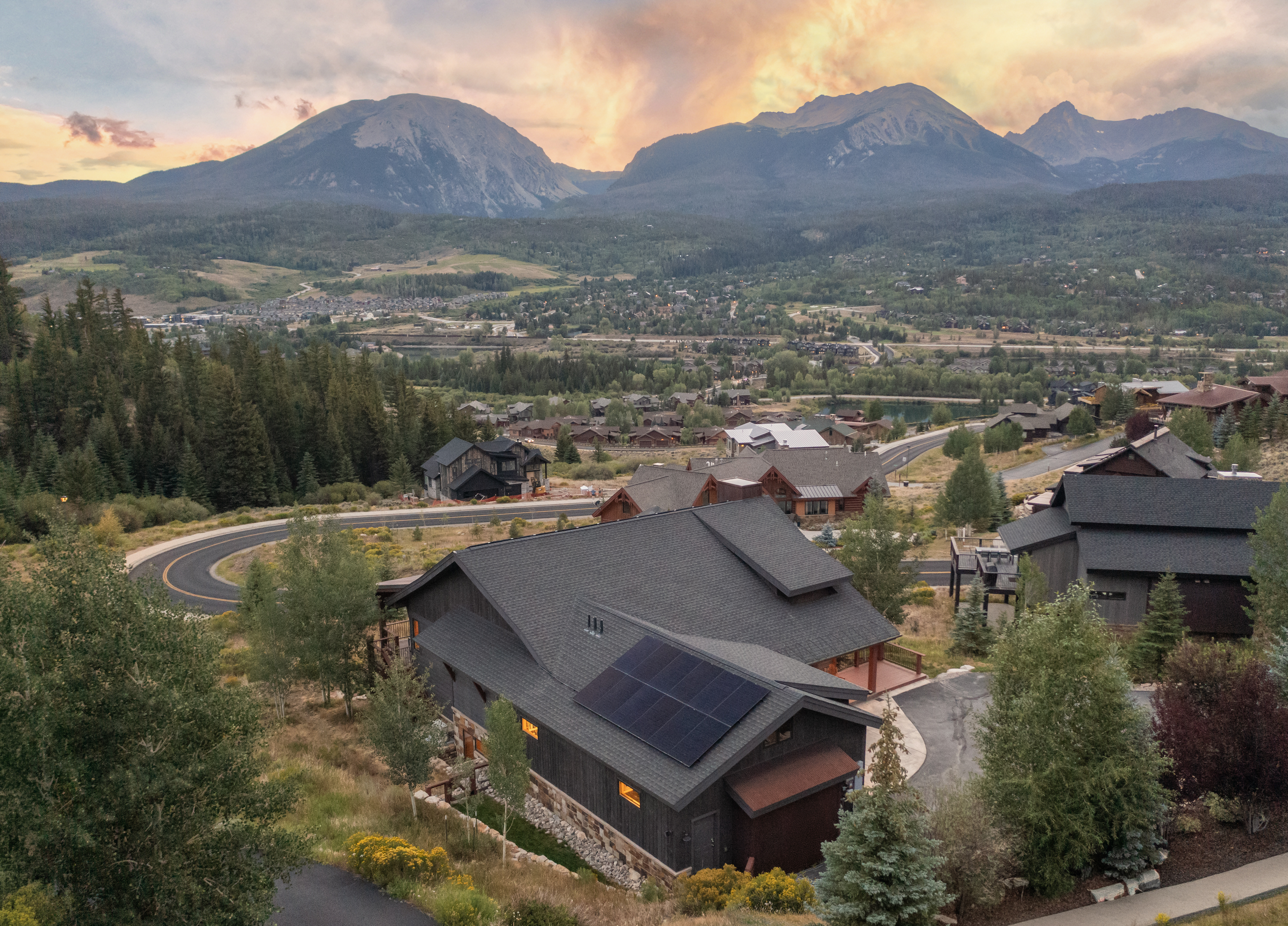 This high-angle aerial shot captures a modern mountain home nestled in a scenic, elevated residential community at sunset. The dark-toned residence features a prominent roof with integrated solar panels, a multi-level design, and a paved driveway, all set against a breathtaking backdrop of rugged, snow-dusted mountain peaks. The surrounding landscape is lush with evergreen trees and winding roads, creating a serene and upscale atmosphere.