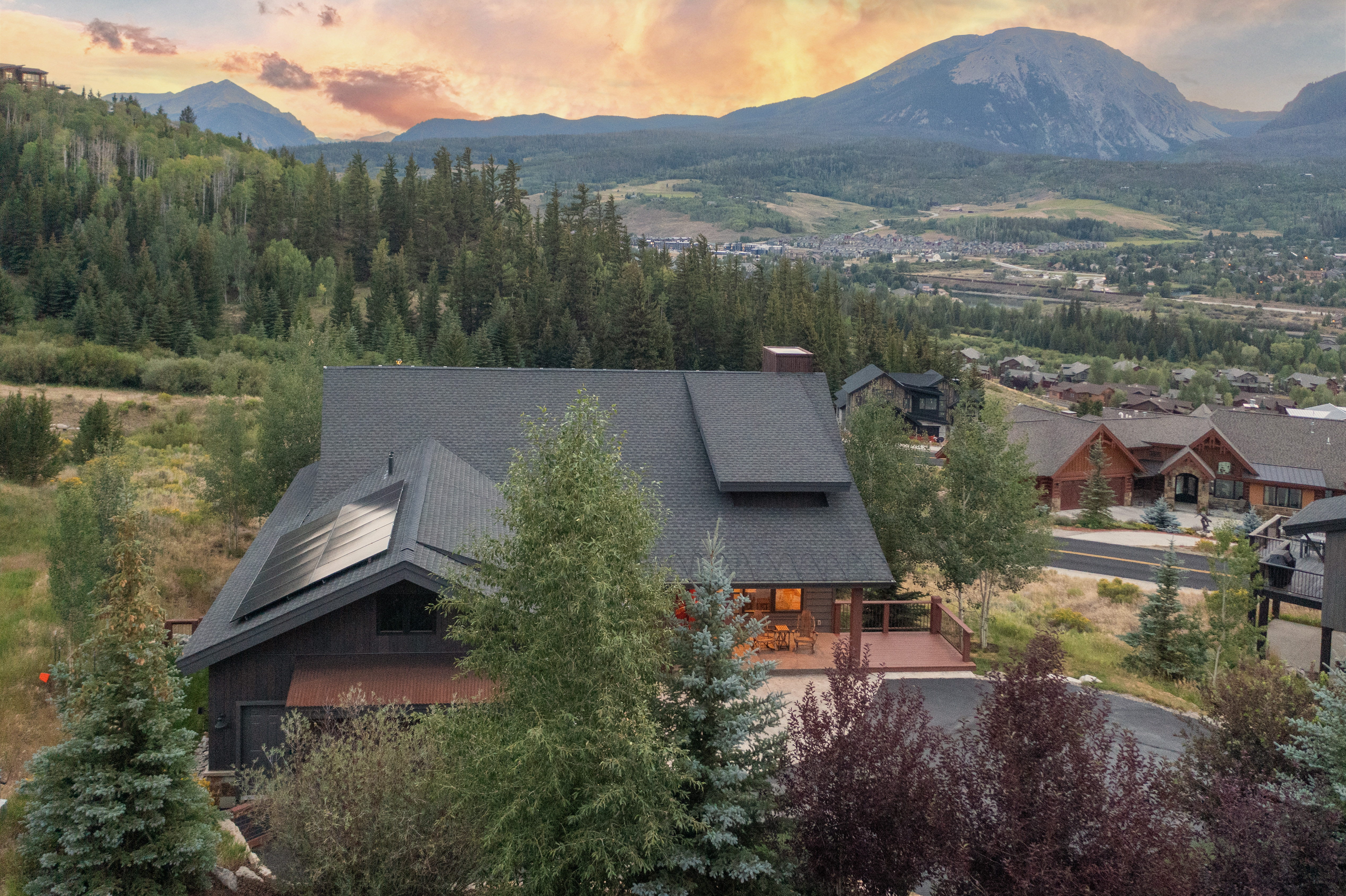 This elevated aerial perspective captures a mountain-style home nestled in a lush, forested landscape at sunset. The house features a dark shingled roof with integrated solar panels, a wooden deck, and a rustic exterior, set against a dramatic backdrop of rolling hills and a prominent mountain peak. The scene conveys a serene, high-altitude residential atmosphere with warm, golden-hour lighting illuminating the surrounding valley.