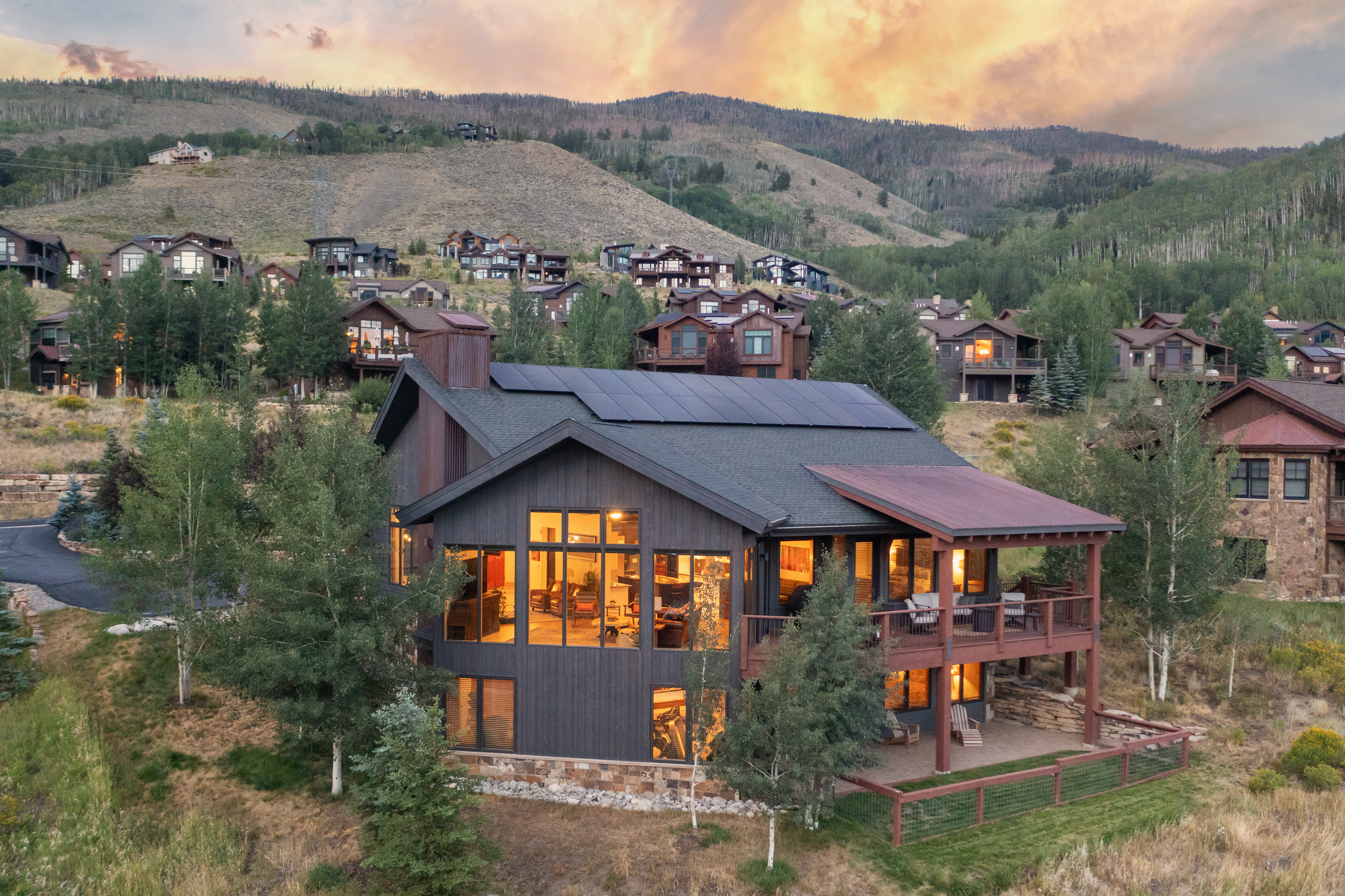 This aerial perspective captures a multi-level mountain home at dusk, set against a backdrop of rolling hills and a vibrant sunset. The residence features dark vertical siding, a prominent roofline with integrated solar panels, and expansive windows that glow warmly from within. A spacious wooden deck extends from the main level, overlooking a natural landscape with mature trees and a fenced yard area.