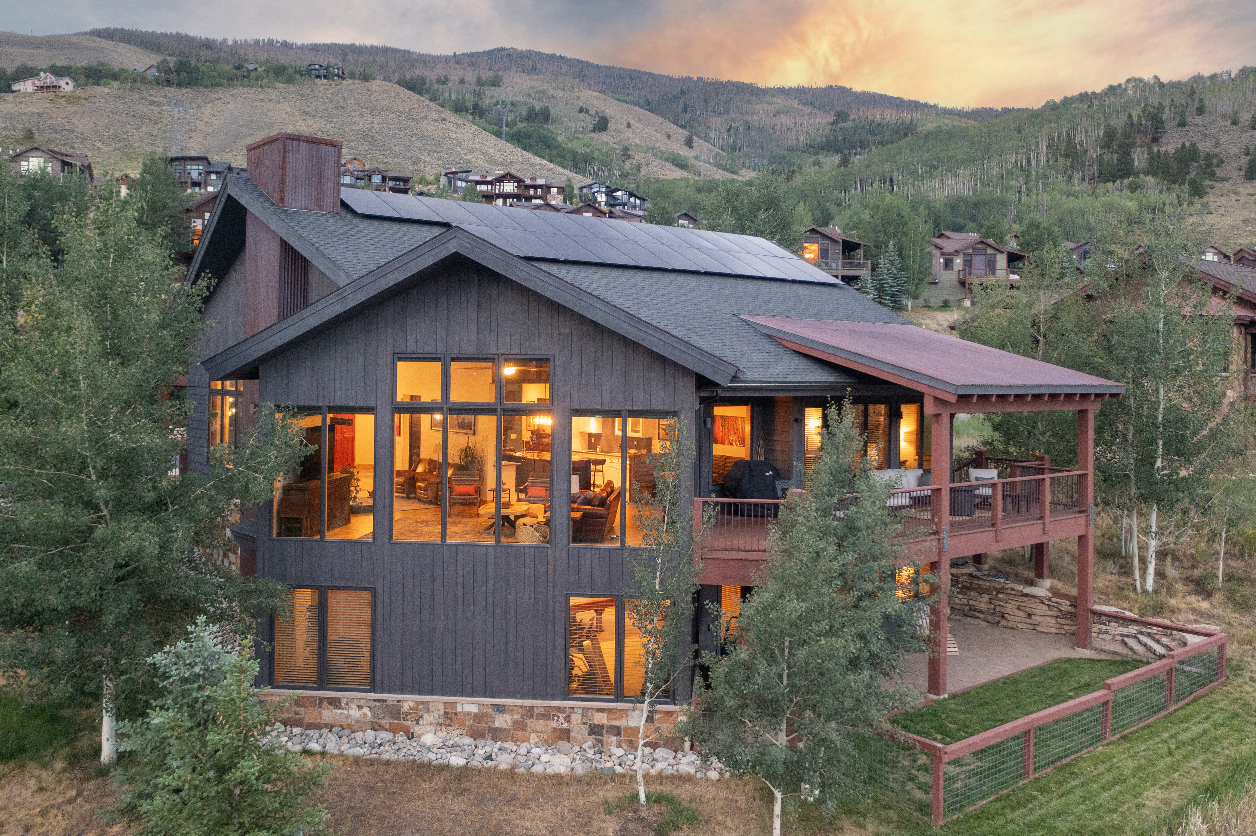 This elevated perspective captures a modern mountain home nestled against a scenic, rolling hillside at dusk. The structure features dark vertical siding, a prominent chimney, and a large deck that overlooks a grassy yard and surrounding aspen trees. The interior living spaces are warmly illuminated, showcasing large windows that create a welcoming glow against the twilight sky.