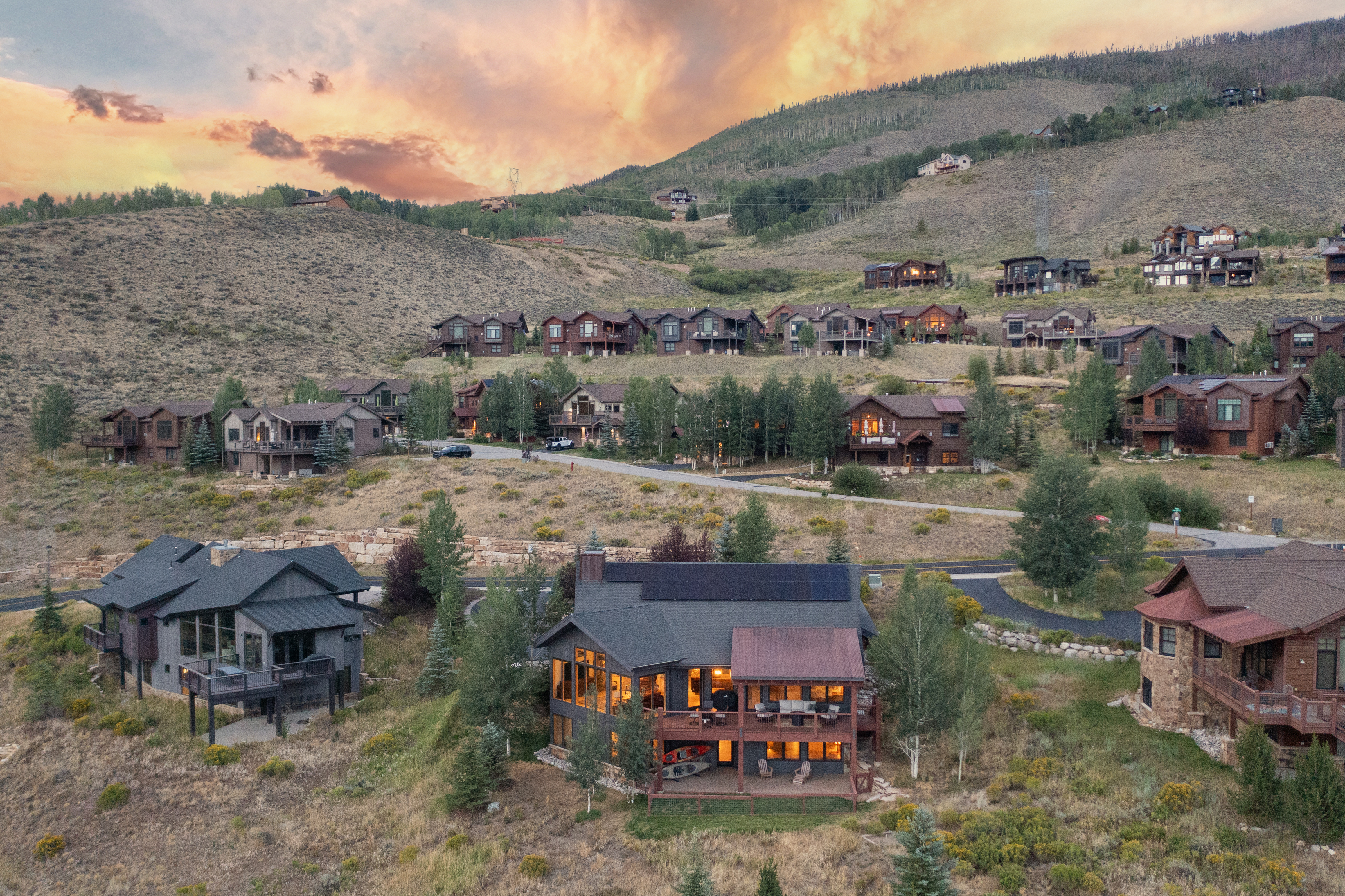 This aerial perspective captures a hillside residential community at sunset, showcasing a collection of multi-story mountain homes nestled into the landscape. The foreground features a prominent residence with a dark shingled roof, solar panels, and expansive windows that glow warmly against the twilight sky. The surrounding terrain is characterized by dry, golden grasses and scattered trees, creating a serene and picturesque mountain retreat atmosphere.