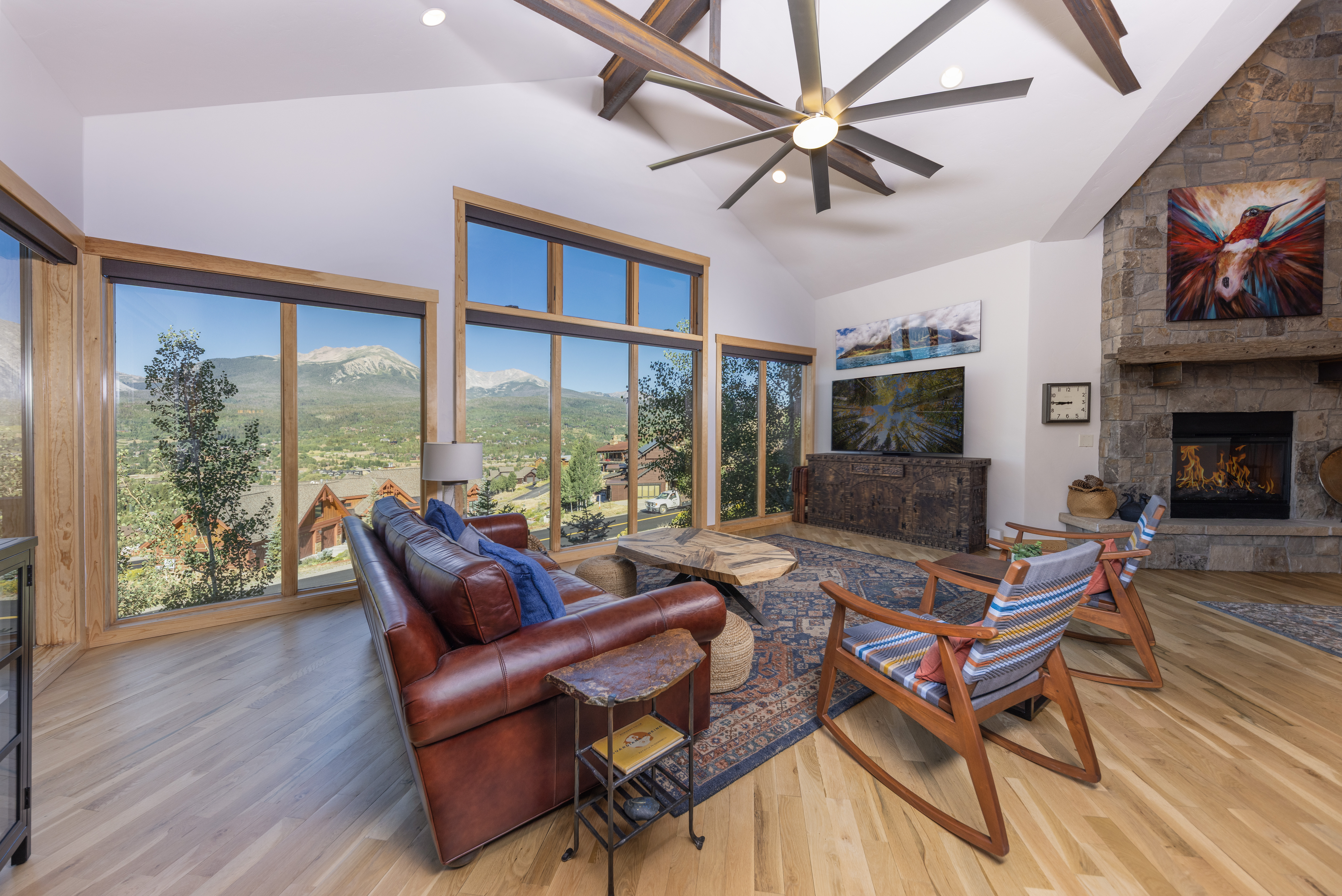 This spacious living room features a vaulted ceiling with exposed wooden beams and a large ceiling fan, creating an airy and rustic atmosphere. A leather sofa and two rocking chairs are arranged around a unique wooden coffee table, all set upon a patterned area rug. The room is anchored by a stone-clad chimney breast with a built-in fireplace, while expansive windows provide panoramic views of the surrounding mountain landscape.