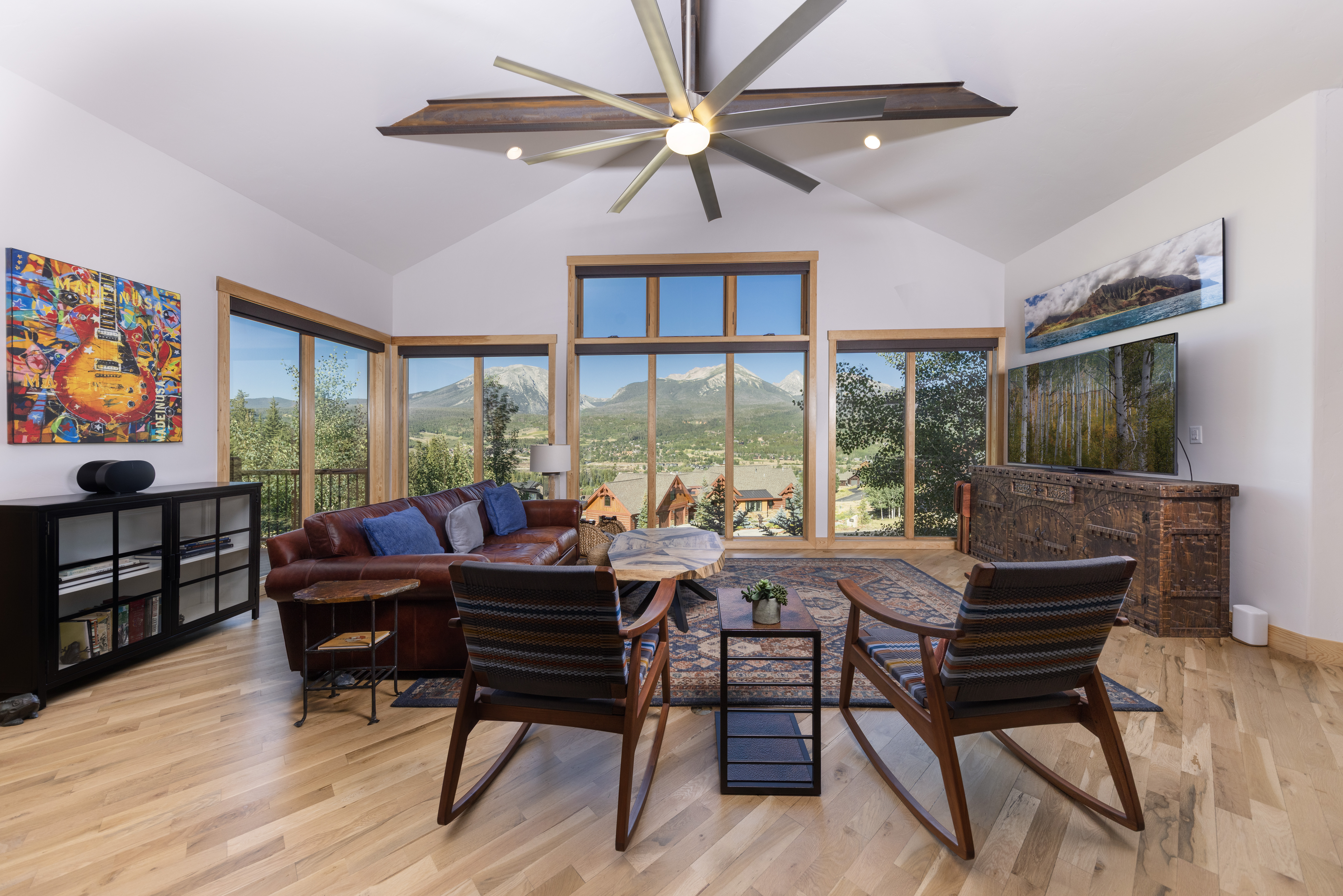 This spacious living room features a vaulted ceiling with a large, modern industrial-style ceiling fan and expansive windows that frame a stunning mountain view. The room is furnished with a rich leather sofa, two wooden rocking chairs, and a unique live-edge coffee table, all arranged on a patterned area rug. A dark wood media console sits to the right, while a black cabinet with glass doors provides additional storage on the left, creating a warm and inviting mountain-lodge aesthetic.