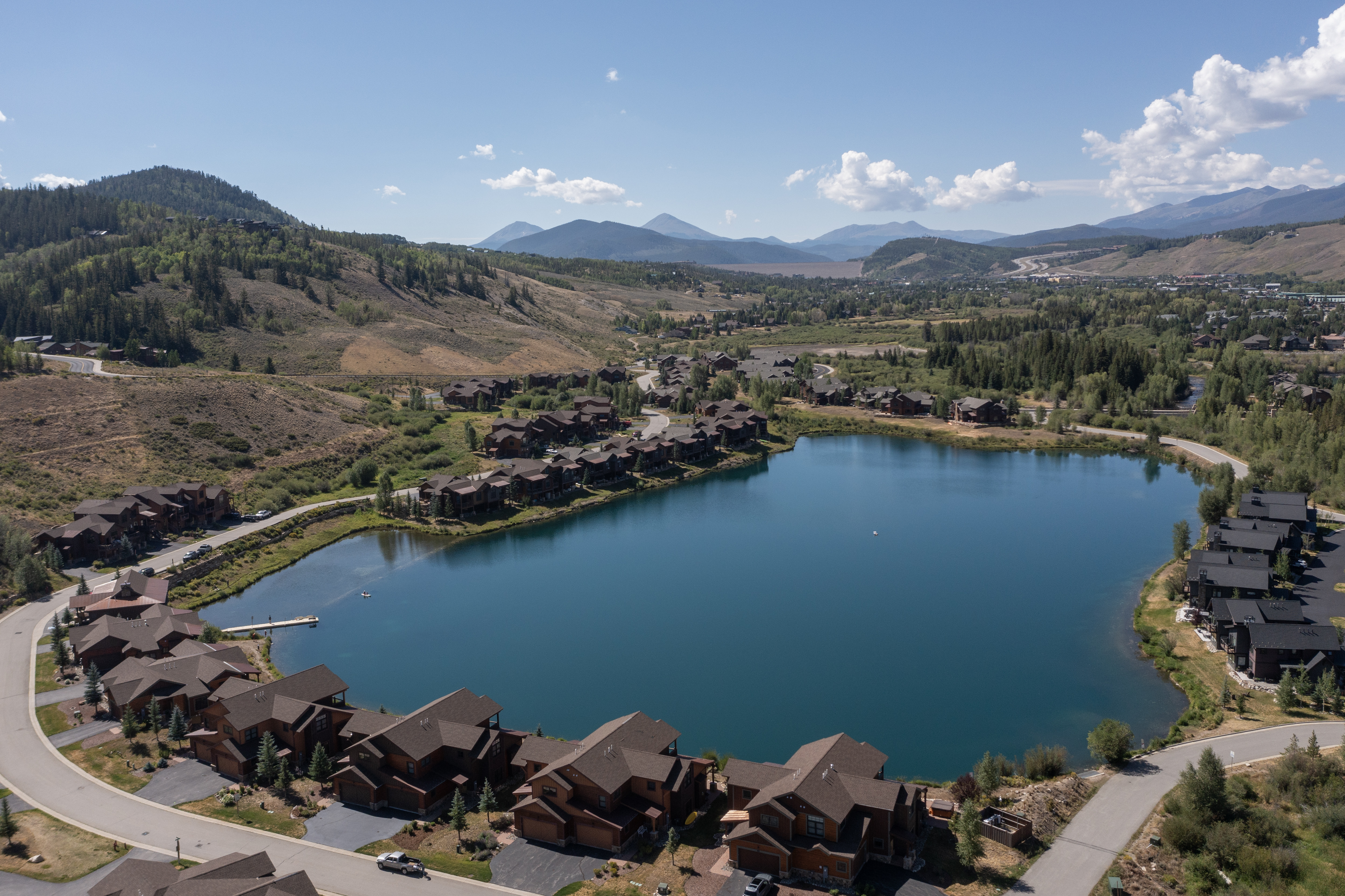 This high-angle aerial view captures a picturesque mountain community centered around a serene, deep blue lake. The landscape features numerous multi-story residential homes with dark-toned roofs and wood-style siding, nestled among rolling hills and dense evergreen forests. In the distance, majestic mountain ranges under a clear blue sky provide a dramatic backdrop, emphasizing the tranquil and scenic nature of this residential development.