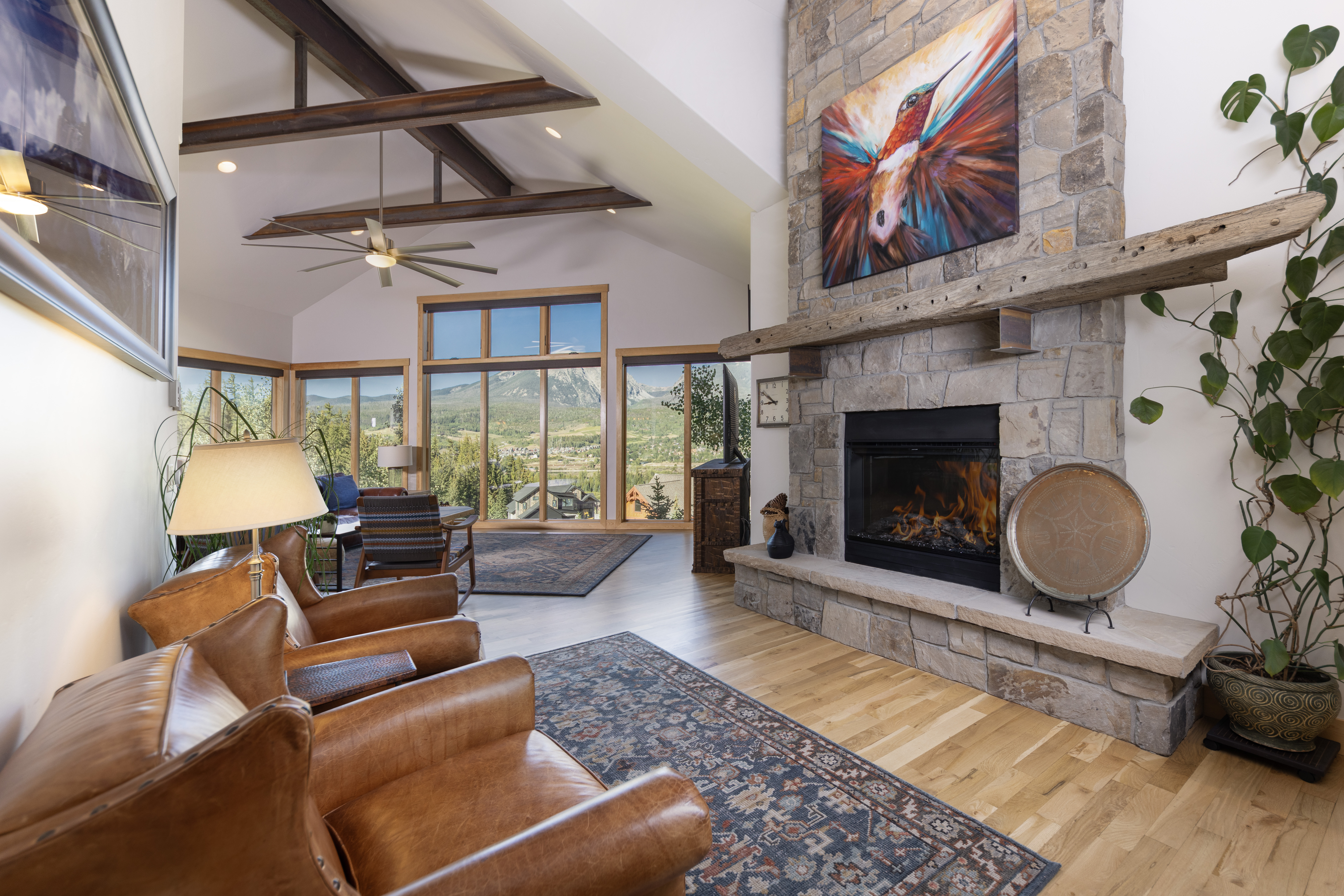 This inviting living room features a prominent stone-clad chimney breast with a vibrant hummingbird painting above the mantel. The space is anchored by two comfortable leather armchairs and a patterned area rug, set against a backdrop of light wood flooring and exposed timber ceiling beams. Large windows provide expansive views of the surrounding mountain landscape, creating a bright and airy atmosphere.