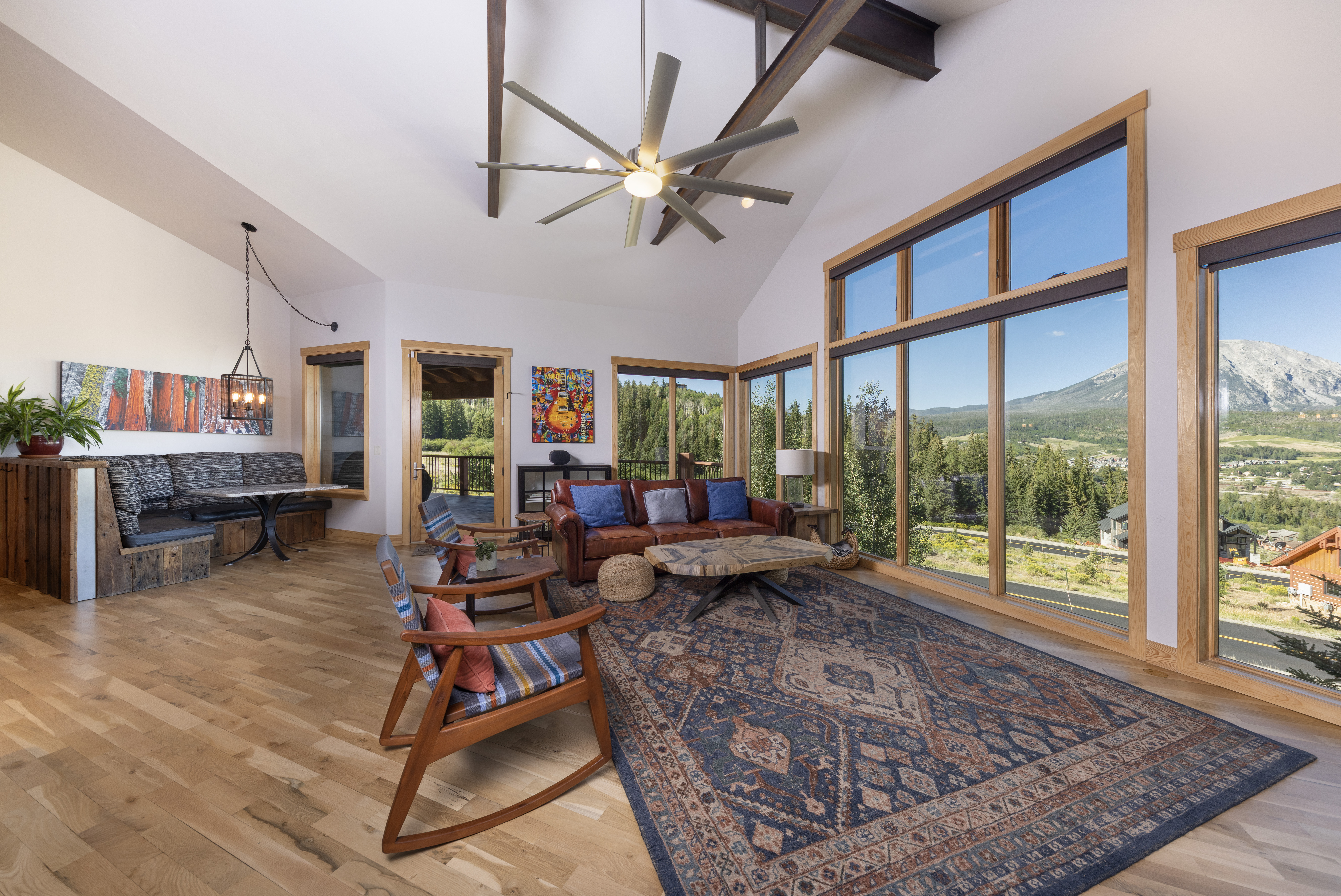This spacious living room features a vaulted ceiling with exposed structural beams and a large ceiling fan, creating an airy, open atmosphere. The room is anchored by a leather sofa and two rocking chairs arranged on a large patterned rug, with a unique live-edge coffee table at the center. Expansive windows provide panoramic views of the surrounding mountains and forest, while a built-in bench seating area with a table offers a cozy nook for dining or relaxing.