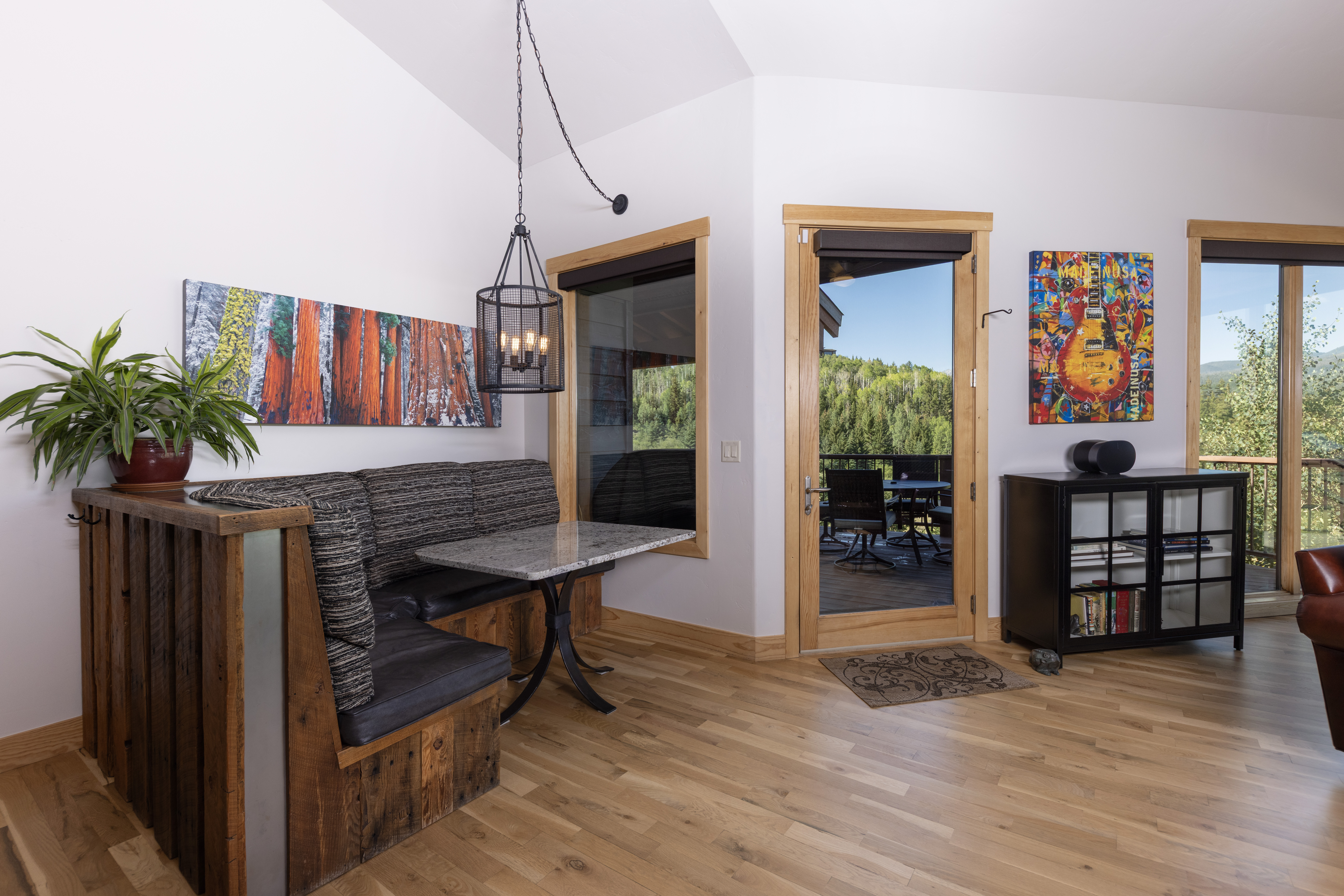 This cozy dining nook features a custom built-in wooden banquette with dark upholstered seating, paired with a stone-topped table and a rustic metal pendant light. The space is accented by vibrant wall art, light wood flooring, and a glass door that opens to a scenic outdoor deck. The overall aesthetic blends rustic charm with a comfortable, inviting atmosphere.