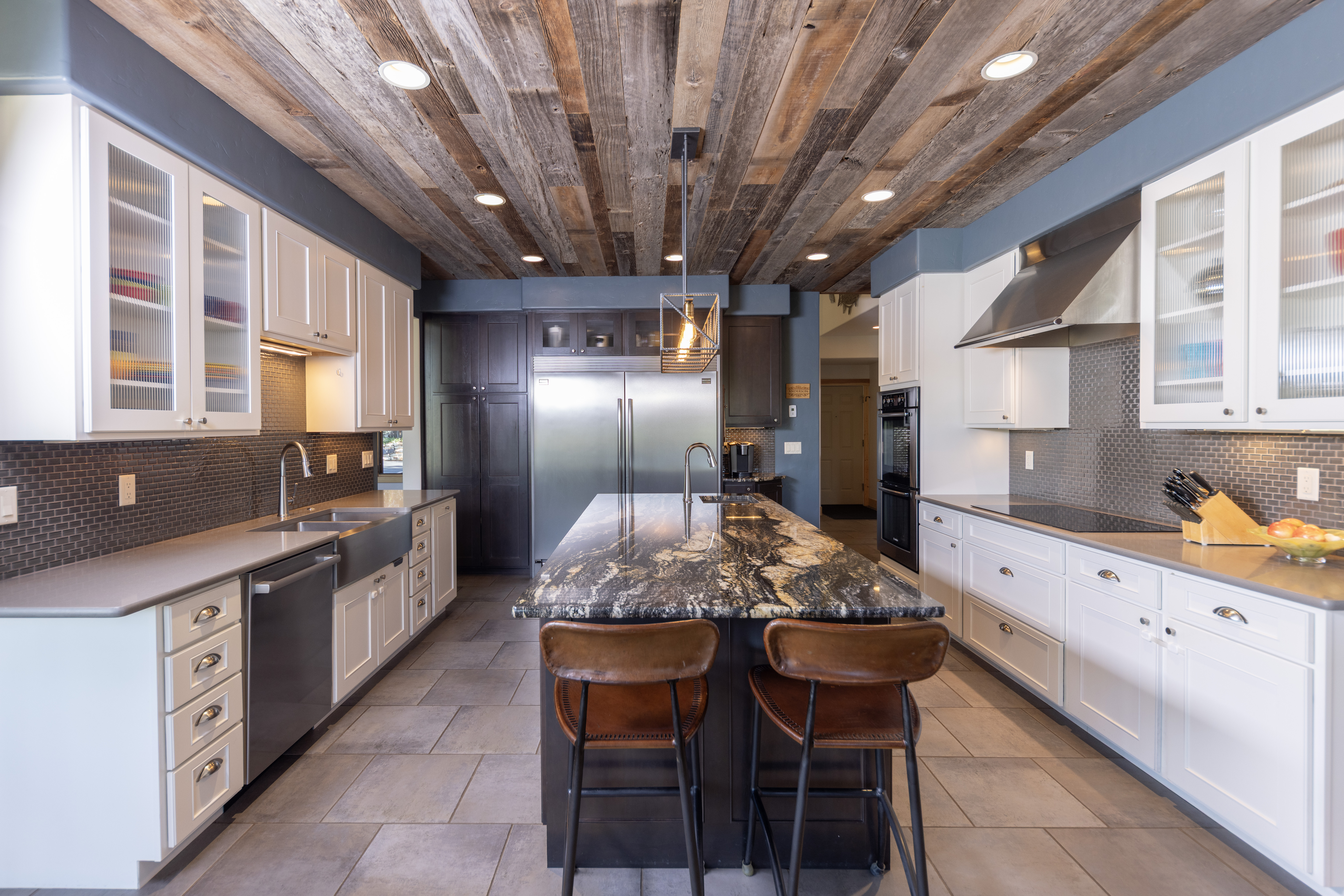 This spacious kitchen features a striking reclaimed wood plank ceiling and a large central island topped with dark, veined stone. The room is equipped with white cabinetry, stainless steel appliances, and a dark tile backsplash, creating a sophisticated contrast. The perspective is a centered, eye-level shot that highlights the symmetrical layout and the warm, inviting atmosphere of the culinary space.