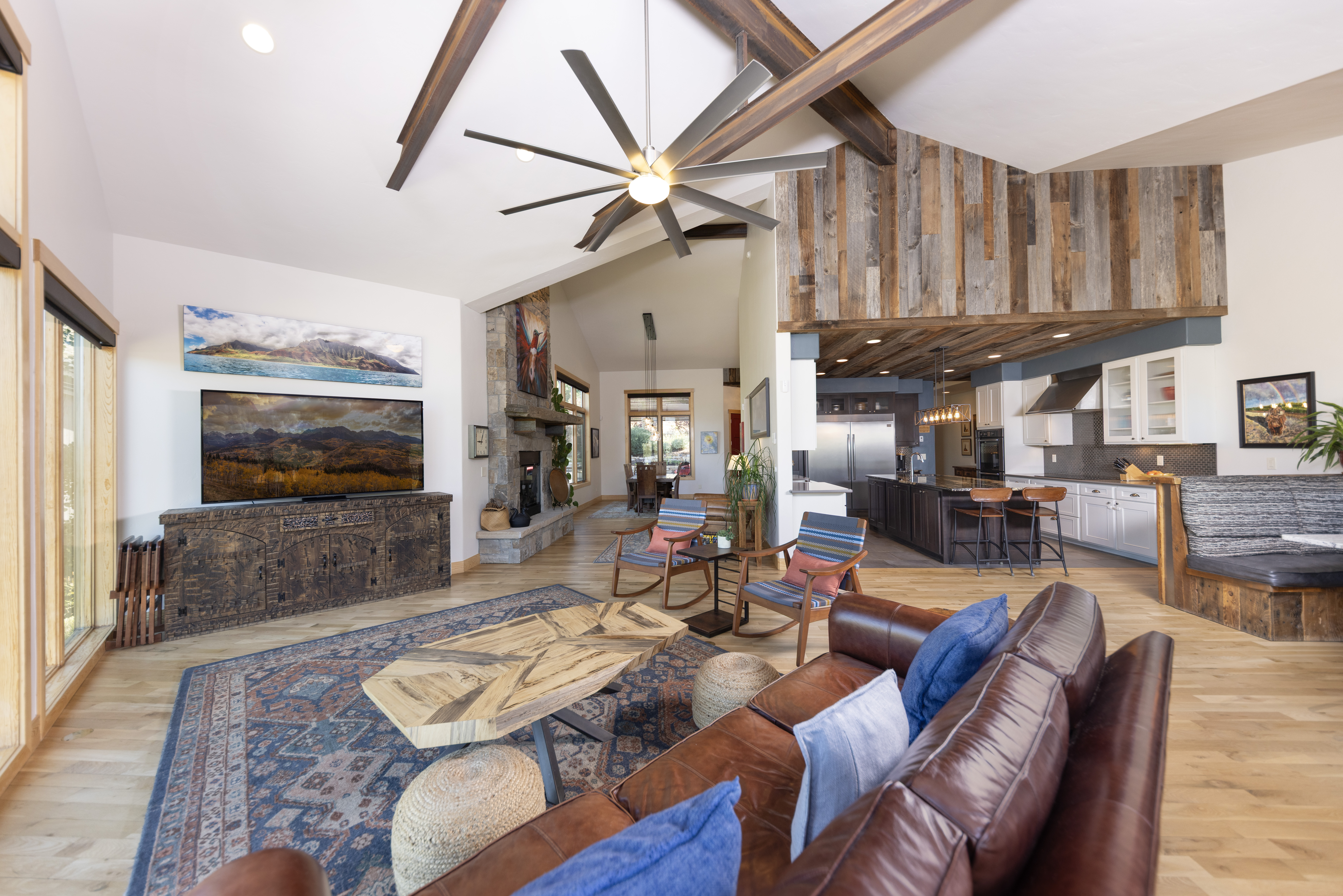 This open-concept living space features a vaulted ceiling with exposed wooden beams and a large, multi-blade ceiling fan. The room is anchored by a leather sofa, a unique geometric coffee table, and a stone-clad chimney breast that serves as a focal point. Beyond the living area, the space flows into a kitchen with a dark-toned island and bar seating, all set against a backdrop of light wood flooring and rustic wood-paneled accents.