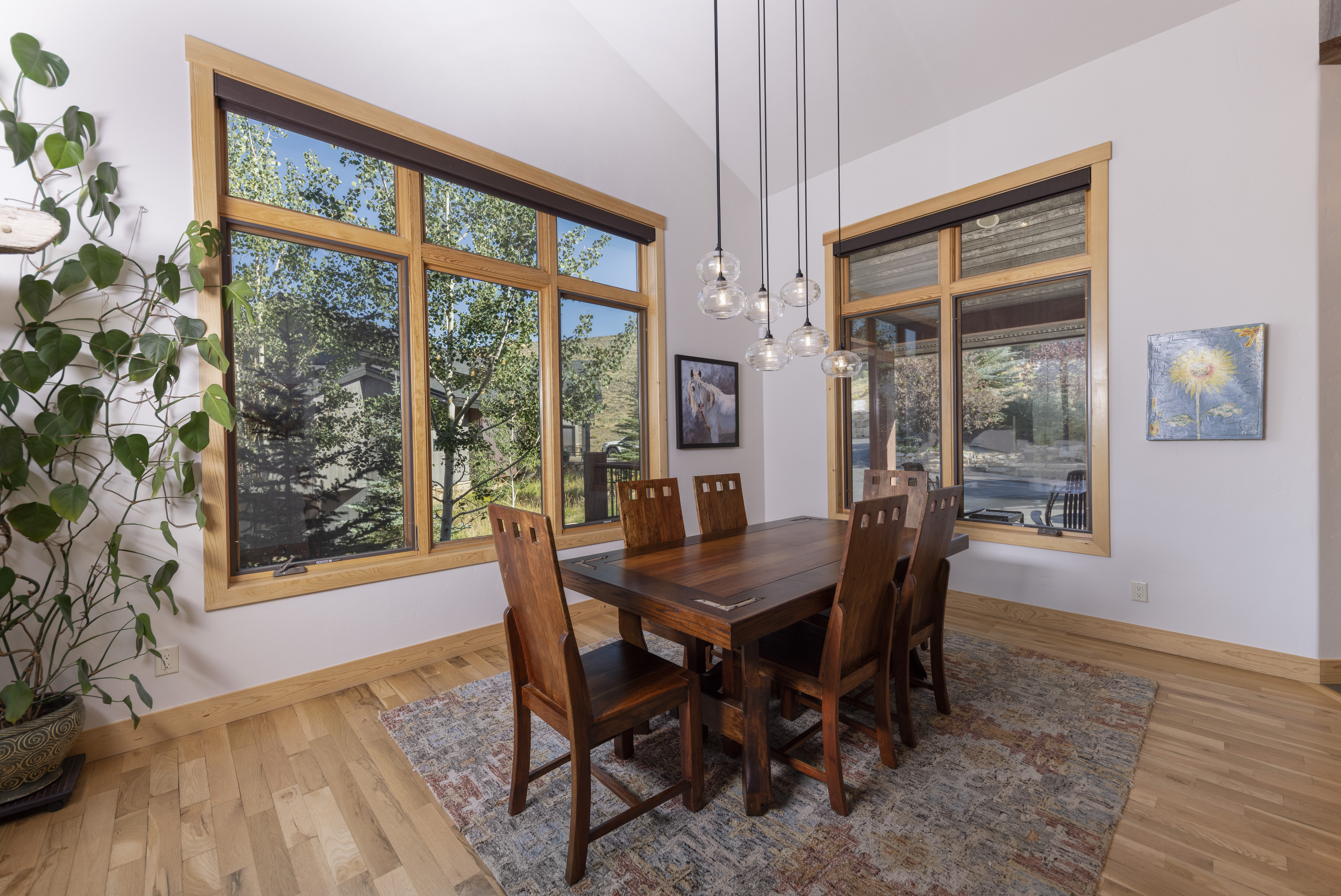 This bright dining room features a dark wood table surrounded by six matching chairs, set atop a textured area rug. A cluster of glass pendant lights hangs above the table, while large windows with light wood trim provide ample natural light and views of the surrounding landscape. The space is accented by a tall potted plant in the corner and two pieces of wall art, creating a warm and inviting atmosphere.