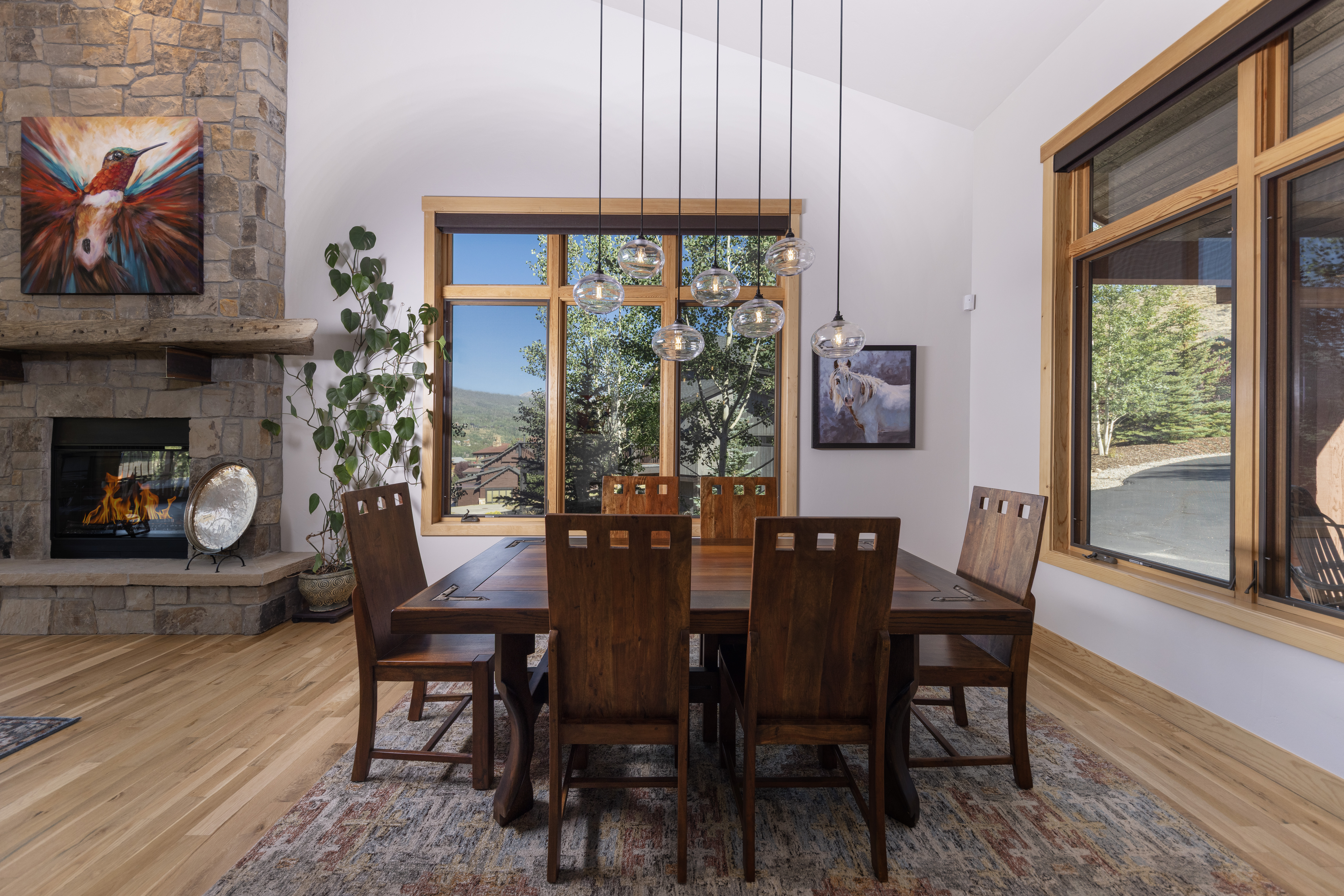 This dining room features a substantial dark wood table surrounded by six matching chairs, set atop a patterned area rug. A stone-clad chimney breast with a rustic timber mantel anchors the space, while a cluster of glass pendant lights hangs above the table. Large windows with light wood frames provide natural light and views of the surrounding landscape, creating a warm and inviting atmosphere.