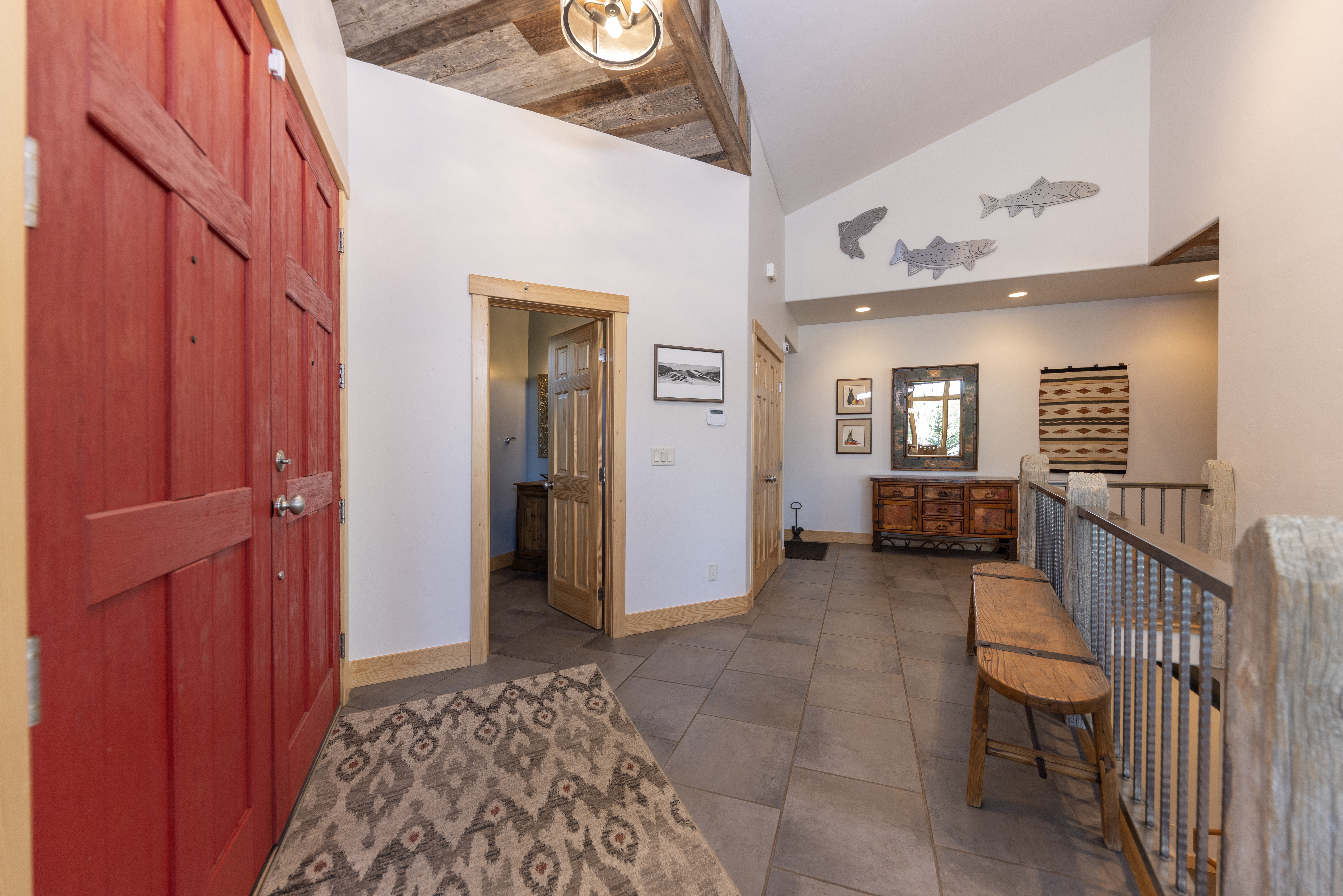 This inviting entryway and hallway feature a rustic aesthetic with a bold red door, patterned area rug, and tile flooring. The space includes a wooden bench, a decorative wooden dresser with a mirror, and wall-mounted fish sculptures that add a unique, lodge-inspired touch. The perspective captures the transition from the entrance into the home, highlighting the vaulted ceiling with exposed wood beams and the open staircase railing.