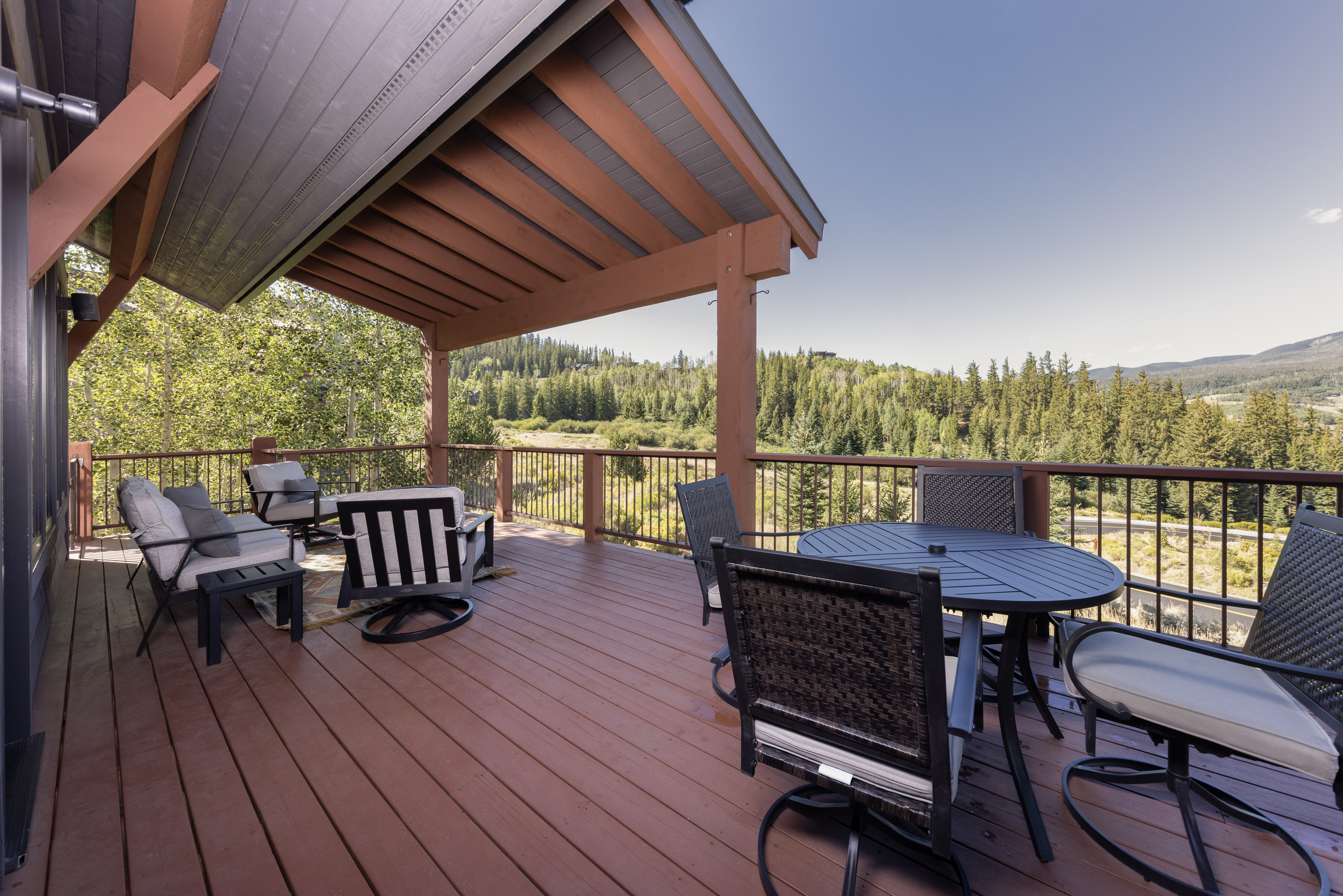 This spacious, elevated wooden deck offers a serene outdoor living space with a covered seating area and an open dining section. The deck is furnished with comfortable lounge chairs and a circular dining set, all overlooking a scenic, tree-lined mountain landscape. The structure features sturdy timber framing and a metal railing, creating a rustic yet inviting atmosphere perfect for enjoying the expansive natural views.