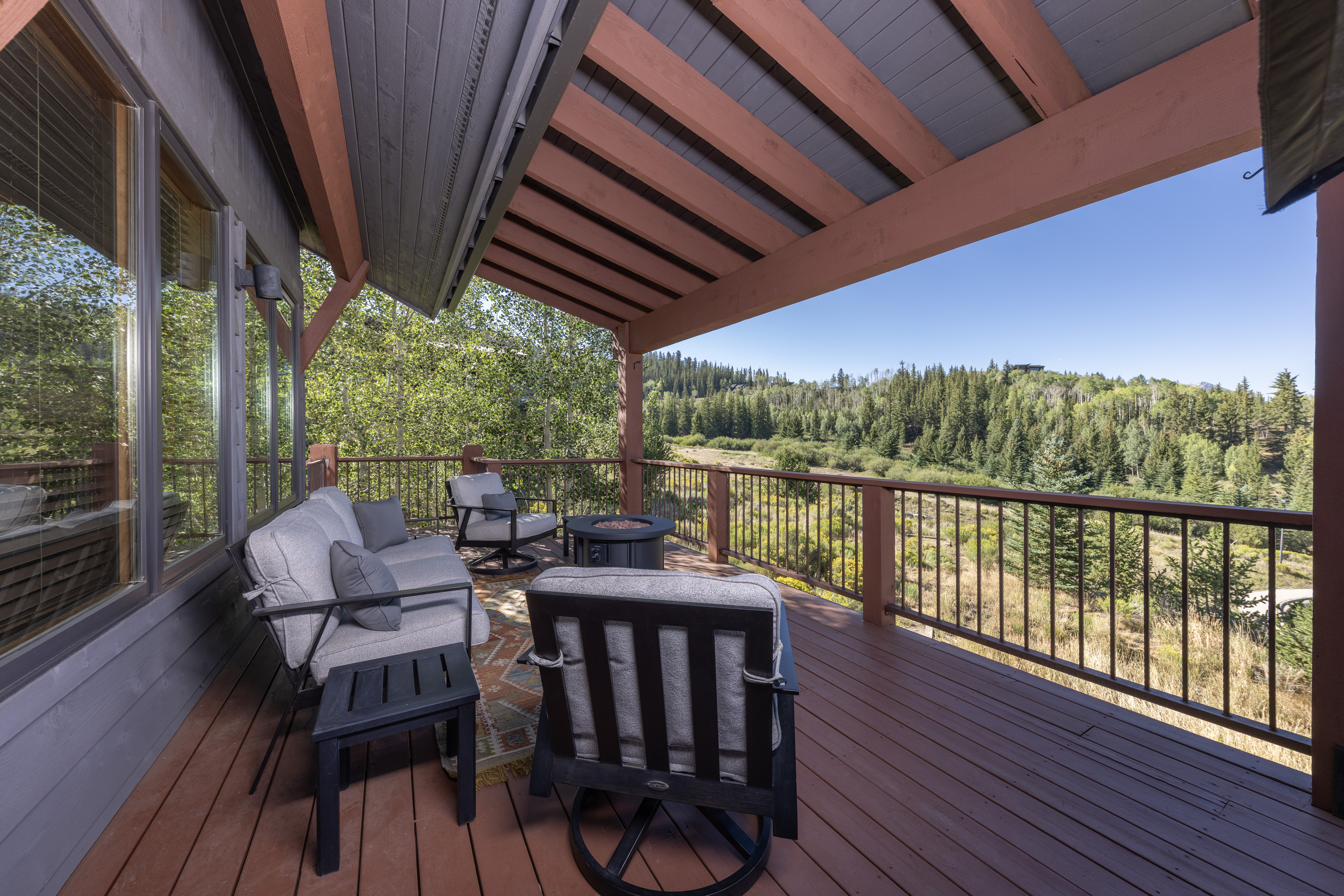 This elevated wooden deck offers a serene outdoor living space with comfortable seating, including a sofa and swivel chairs arranged around a central fire pit. The deck features a dark metal railing and is partially covered by a timber-framed roof structure, providing a perfect vantage point for the expansive, lush forest views beyond. The overall atmosphere is peaceful and inviting, highlighting the home's connection to the surrounding natural landscape.