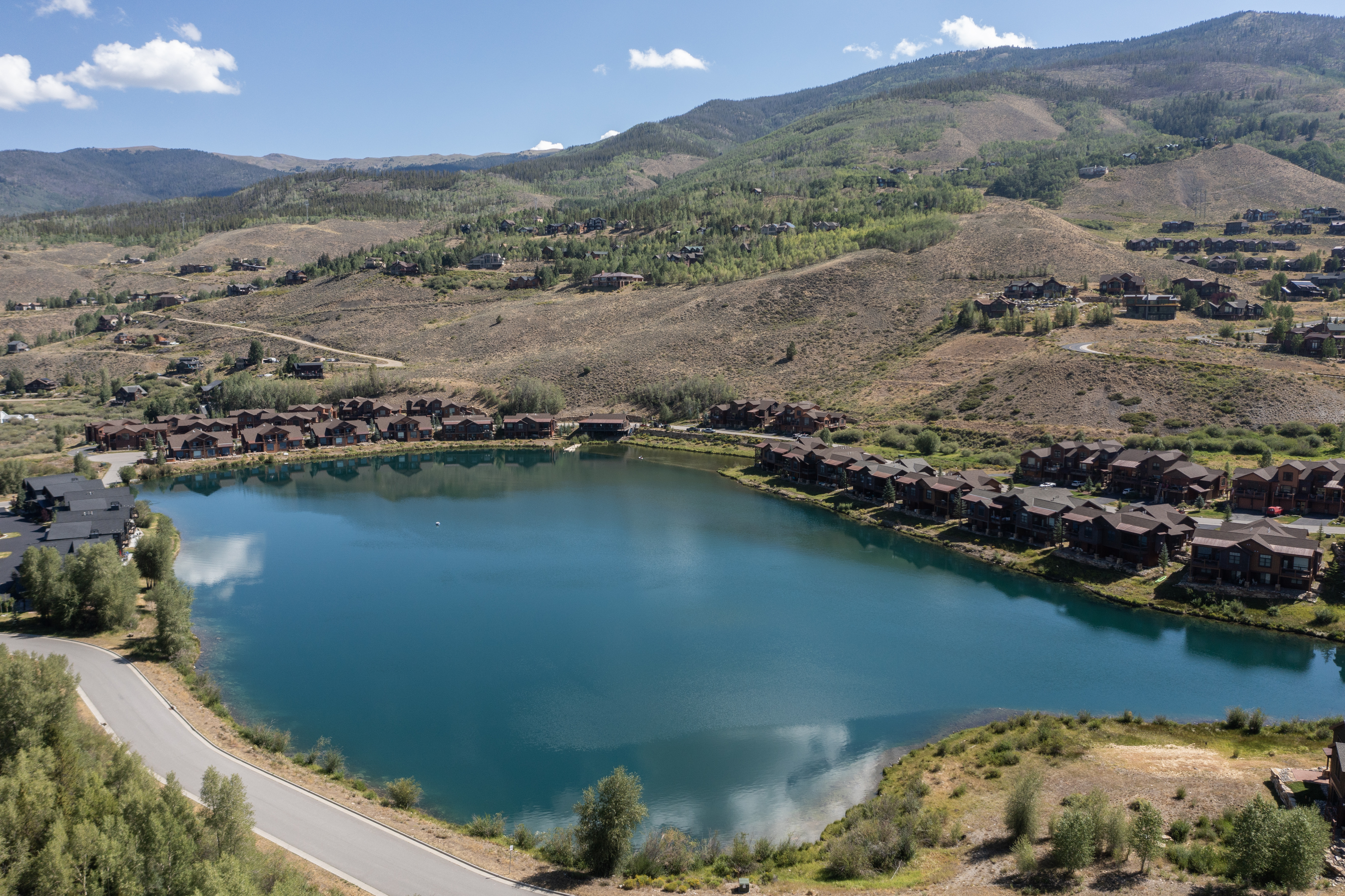 This high-angle aerial view captures a serene mountain community centered around a vibrant blue lake. The landscape features clusters of rustic, multi-story residential buildings with dark wood siding and gabled roofs, nestled into the rolling, arid hillsides. A winding paved road leads into the development, providing a sense of scale and accessibility to this tranquil, scenic retreat.