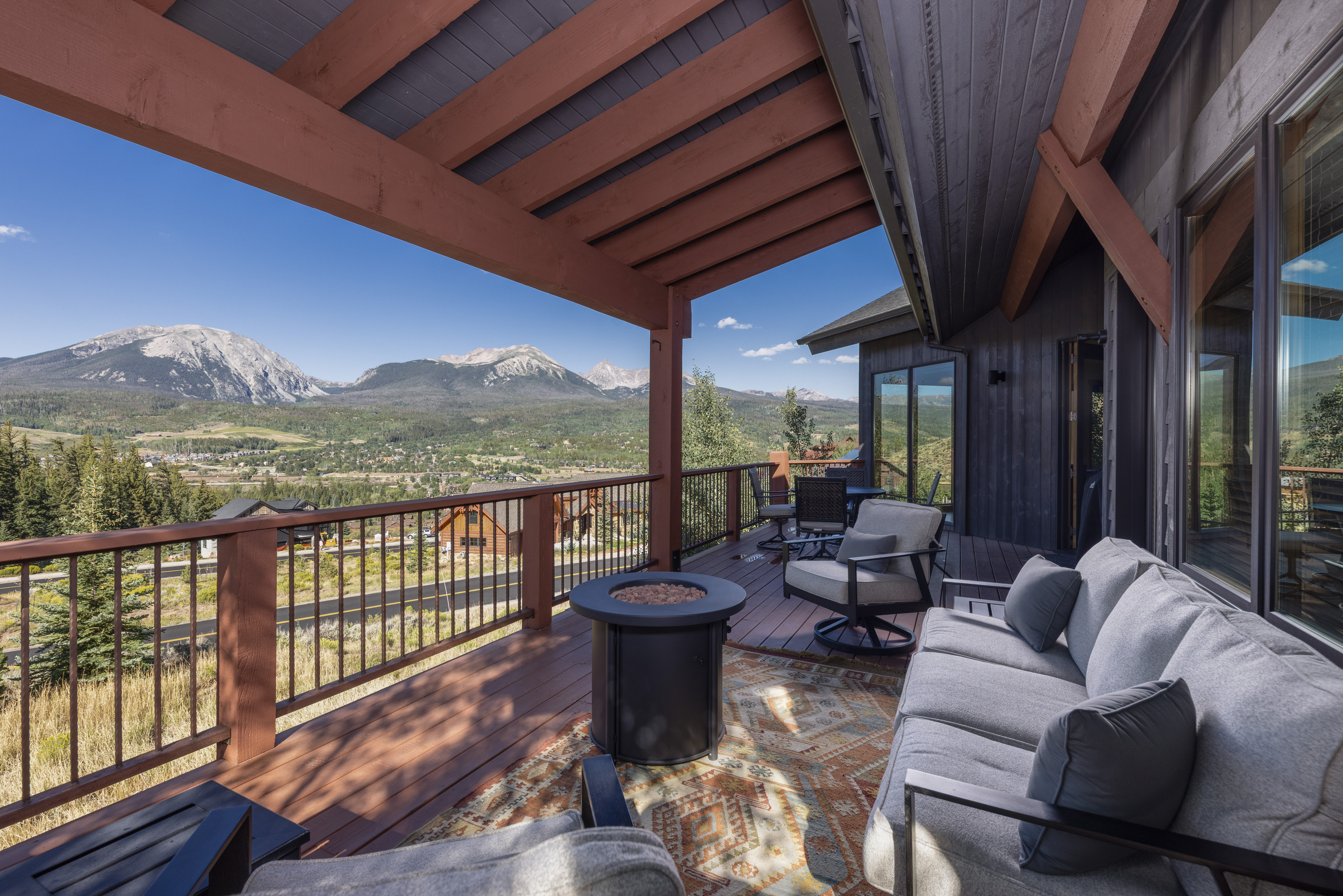 This expansive wooden deck offers a stunning panoramic view of a mountain valley and town below. The space is furnished with a comfortable outdoor sofa, swivel armchairs, and a central fire pit, all set atop a patterned area rug. The covered deck features exposed timber beams and dark-toned siding, creating a rustic yet sophisticated atmosphere perfect for outdoor relaxation.