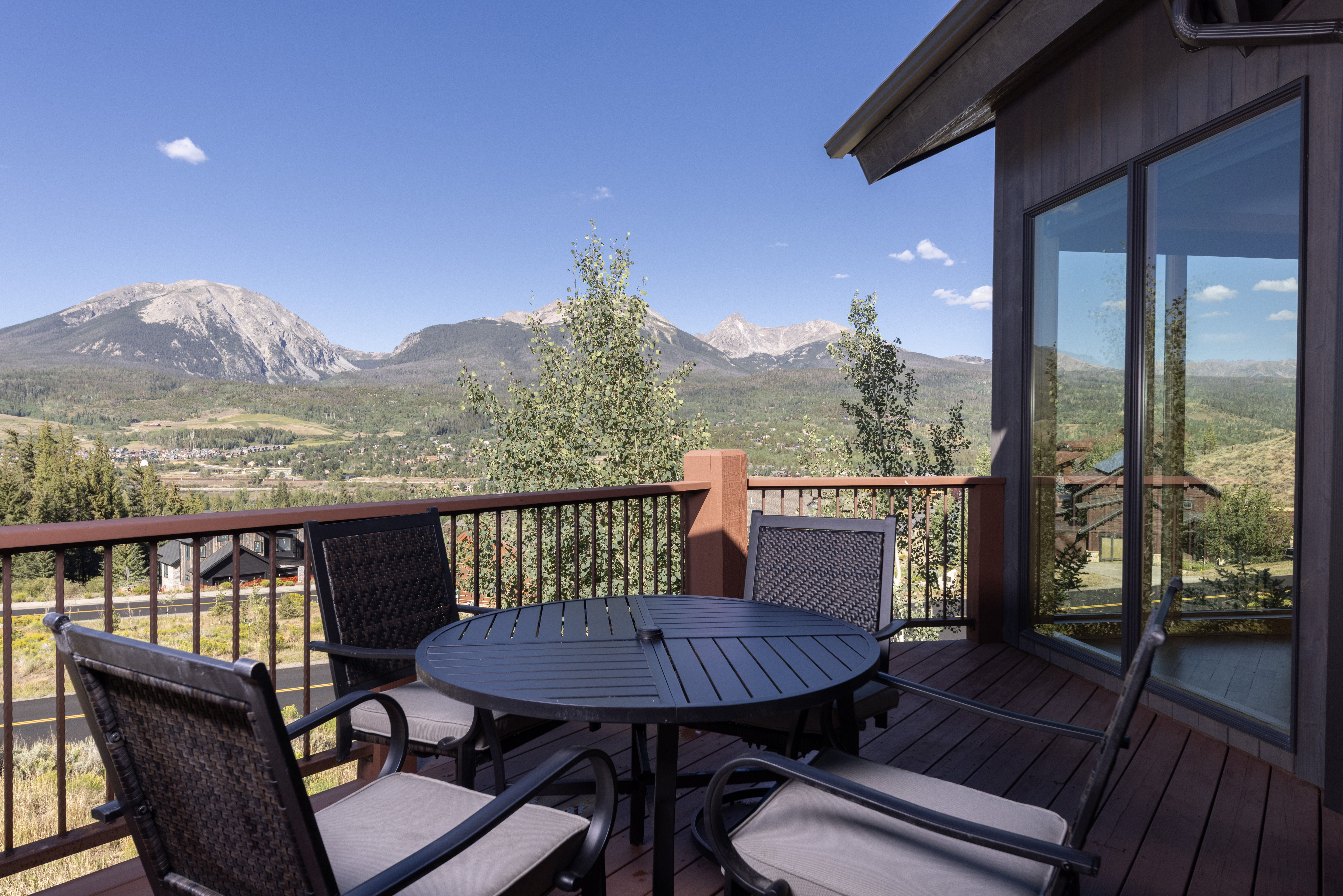 This elevated deck offers a stunning panoramic view of a mountain range and a valley town below. The space is furnished with a circular metal table and four matching chairs with cushioned seats, providing an ideal spot for outdoor dining or relaxation. Large glass panels on the adjacent wall reflect the surrounding landscape, enhancing the connection between the indoor and outdoor living areas.