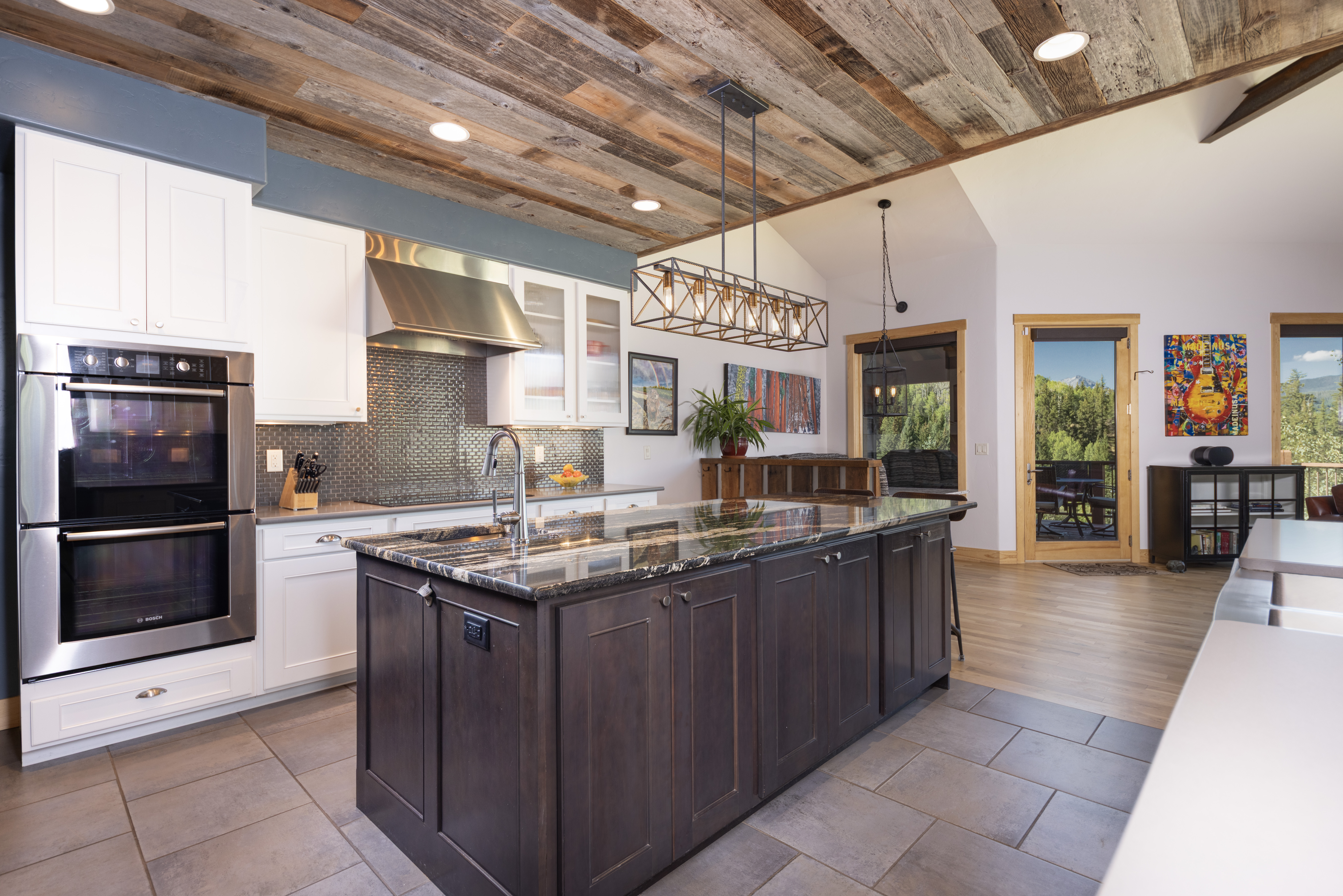 This spacious kitchen features a large dark-toned central island with a stone countertop, contrasting against white perimeter cabinetry and a metallic tile backsplash. The room is defined by a rustic wood-plank ceiling and a prominent rectangular pendant light fixture hanging above the island. A double wall oven is integrated into the cabinetry, and the open layout flows toward a doorway leading to an outdoor deck with mountain views.