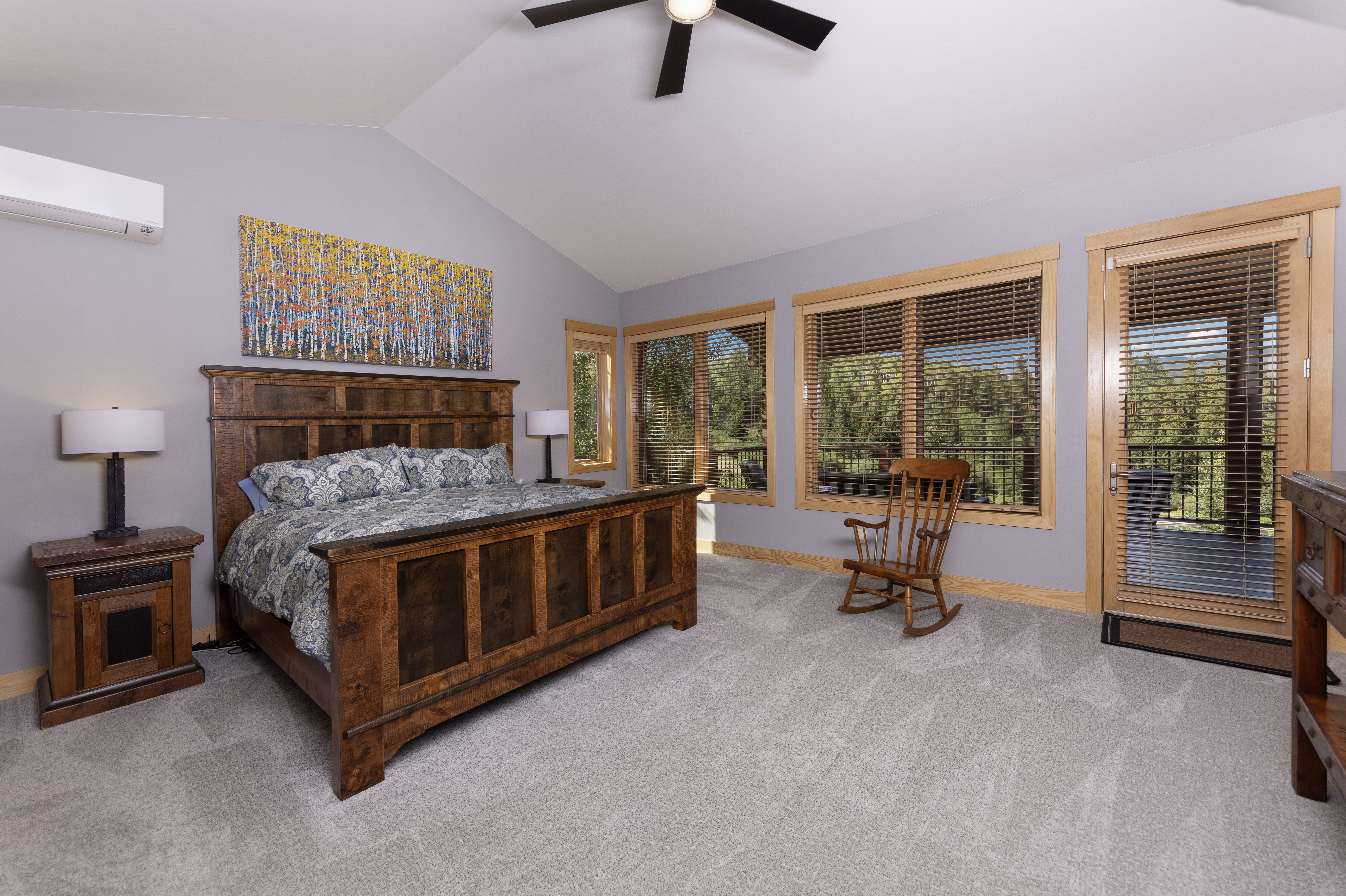 This spacious primary bedroom features a large, rustic wooden bed frame as the centerpiece, complemented by matching nightstands and a rocking chair. The room is bathed in natural light from several windows and a glass-paneled door that opens to an outdoor deck, offering a serene view of the surrounding trees. A vibrant, textured painting of an aspen forest hangs above the bed, adding a pop of color to the neutral gray walls and carpeted floor.