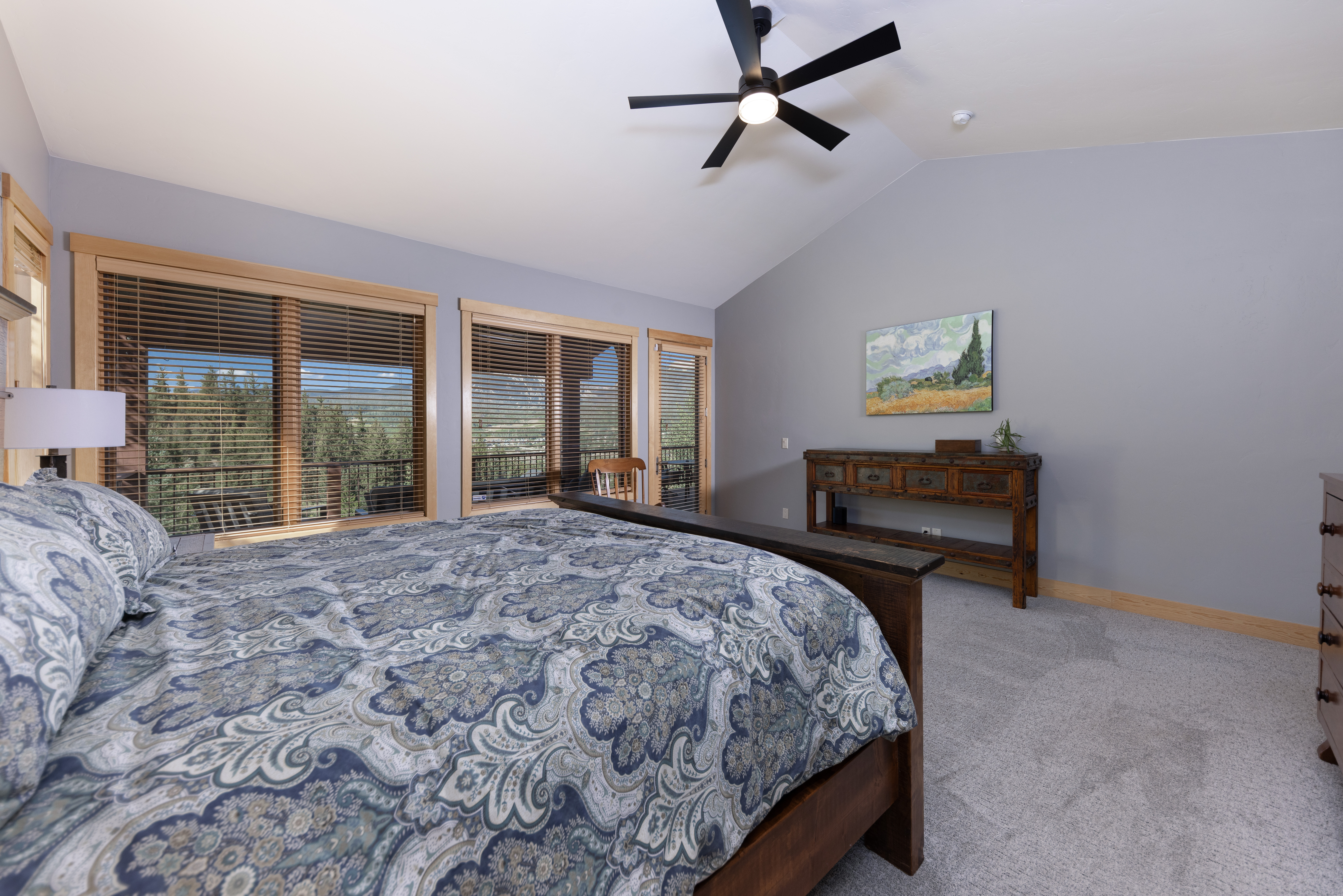 This primary bedroom features a vaulted ceiling with a central black ceiling fan and light fixture. A large bed with a patterned blue and white comforter dominates the foreground, while a rustic wooden console table sits against the gray wall, topped with a landscape painting. Large windows with wooden frames and blinds offer a view of the surrounding trees and mountains, creating a bright and serene atmosphere.