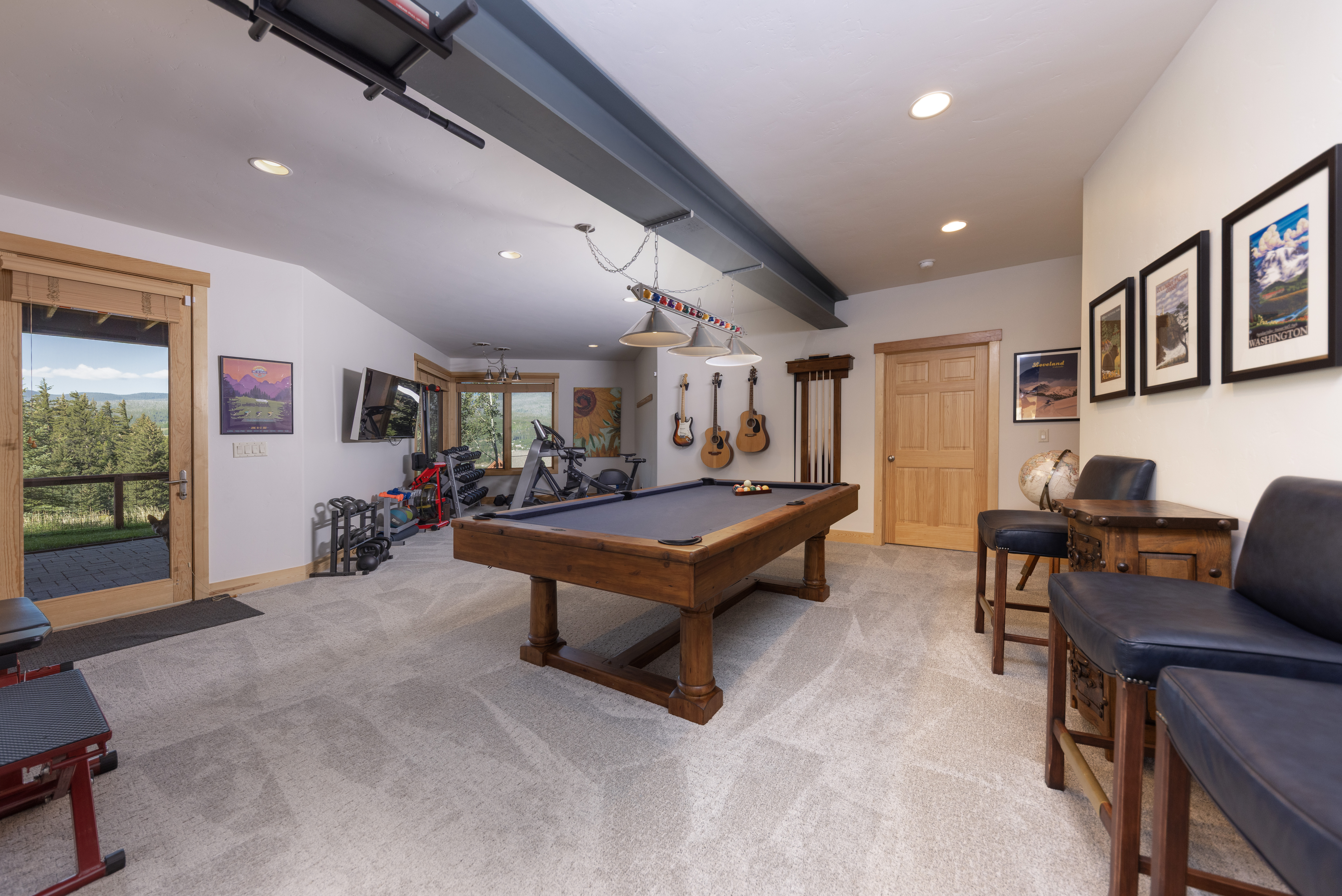 This spacious, multi-functional basement features a central pool table illuminated by hanging pendant lights, creating a dedicated game area. The room seamlessly integrates a home gym setup with various weights and equipment, alongside a wall display of acoustic guitars. With neutral carpeting, light-colored walls, and a glass door leading to an outdoor view, the space offers a versatile and inviting recreational environment.
