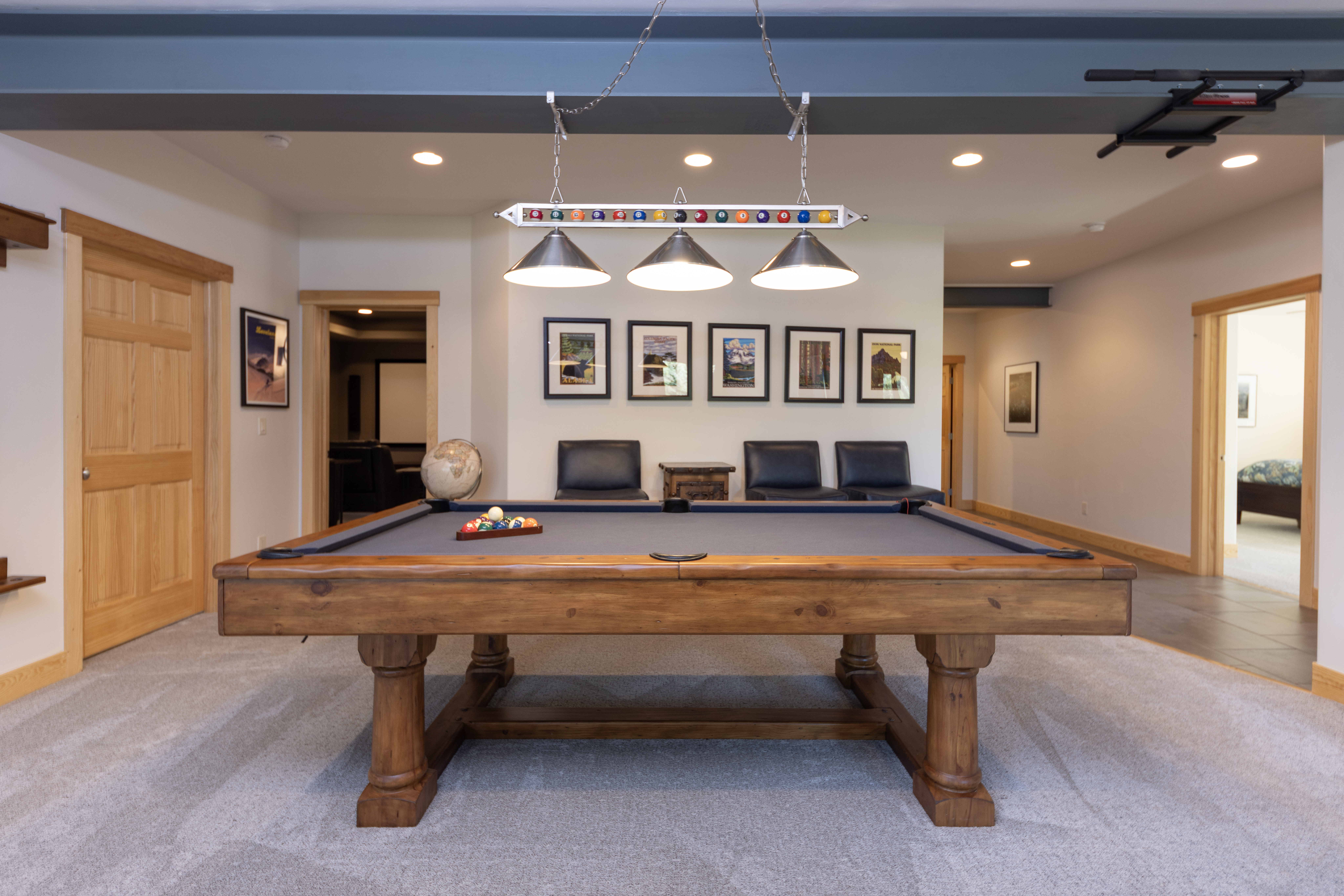 This spacious basement recreation area features a prominent wooden pool table as the central focal point, illuminated by a multi-light billiard fixture. The room is decorated with a series of framed landscape prints on the wall, accompanied by a row of dark leather chairs and a small side table. The neutral carpeted flooring and light-colored walls create a comfortable, inviting atmosphere suitable for entertainment and leisure.