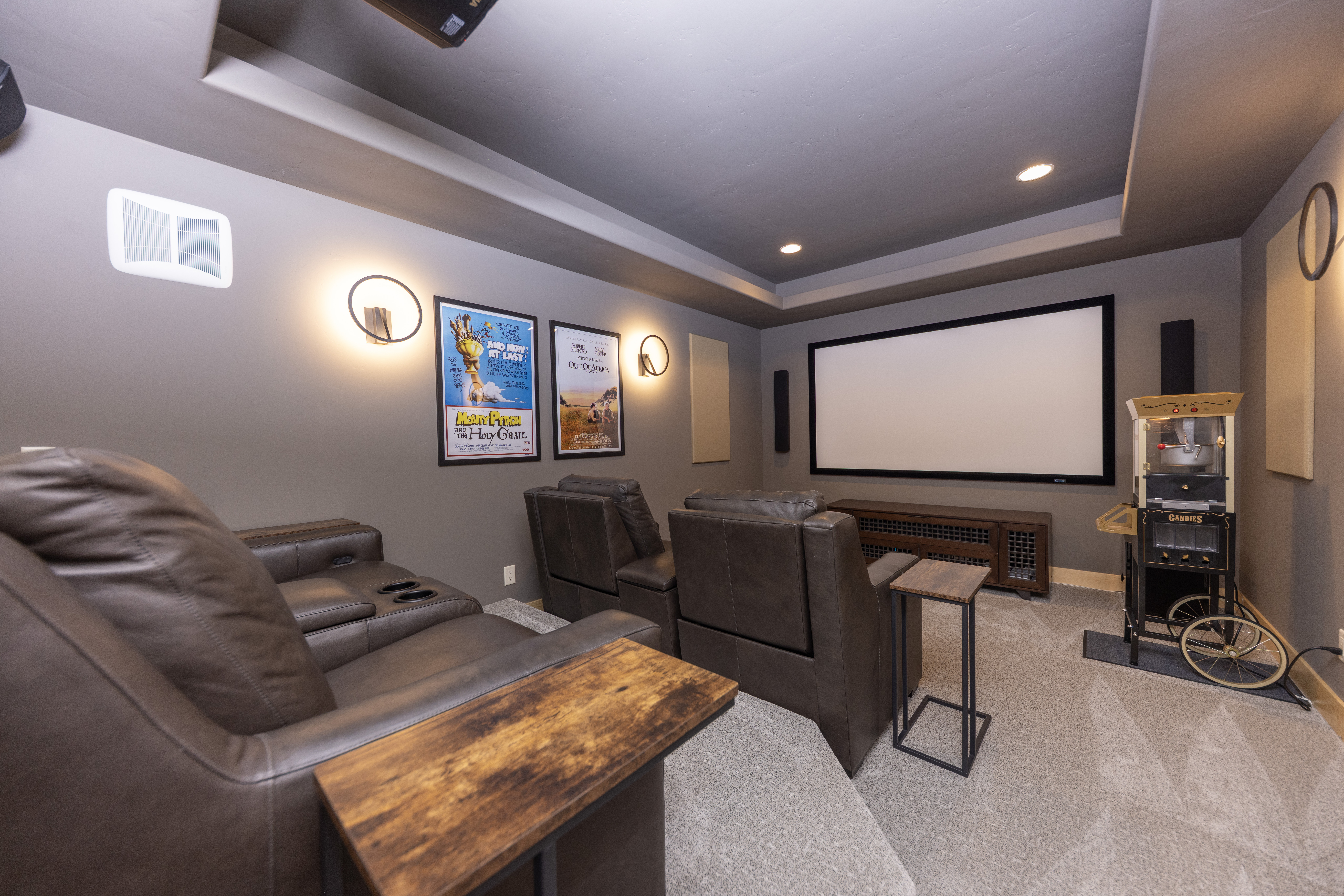 This dedicated home theater room features tiered seating with comfortable brown leather recliners and two side tables. The space is equipped with a large projection screen, a media console, and a vintage-style popcorn machine, creating an authentic cinematic atmosphere. Wall-mounted movie posters and circular sconce lighting enhance the cozy, immersive design of this entertainment area.