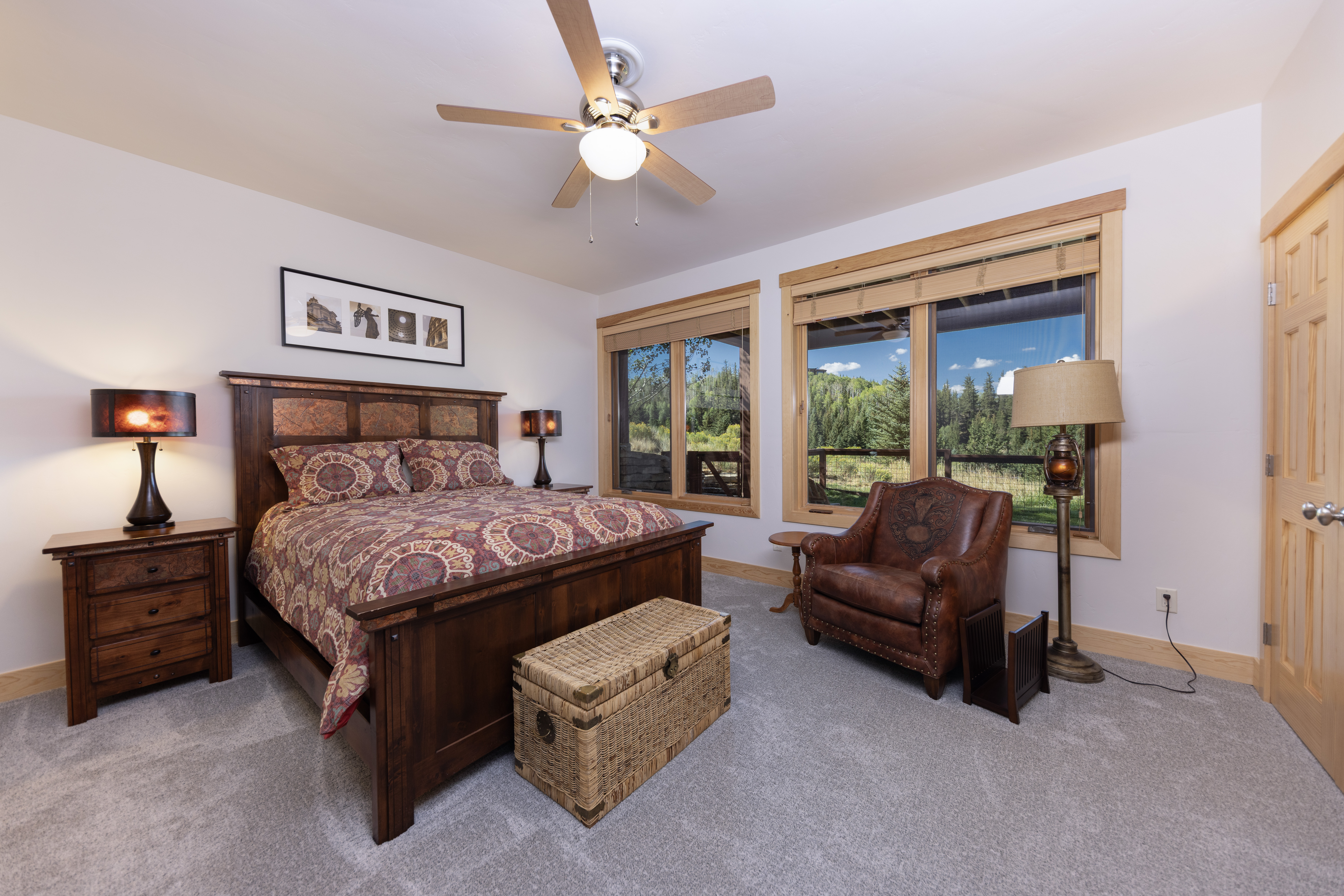 This cozy bedroom features a rustic wooden bed frame with a patterned quilt and a matching wooden nightstand topped with a lamp. A leather armchair sits near large windows that provide a scenic view of the surrounding forest, while a woven trunk rests at the foot of the bed. The room is finished with neutral walls, light-colored carpeting, and a ceiling fan, creating a warm and inviting atmosphere.