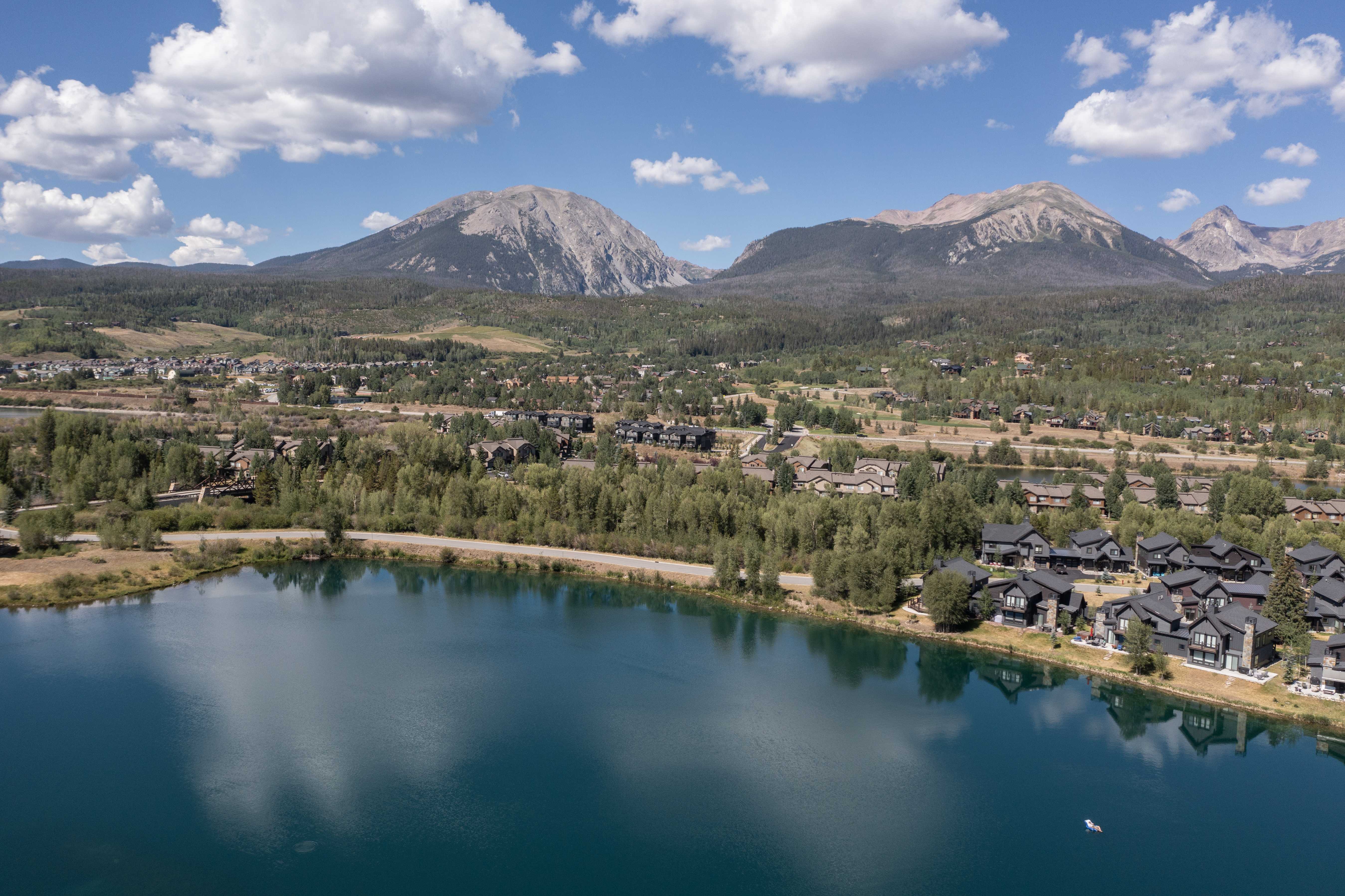 This expansive aerial view captures a serene mountain community nestled beside a vibrant, deep-blue lake. The scene features a cluster of modern, dark-toned residential homes situated along the water's edge, set against a backdrop of lush green forests and towering, rugged mountain peaks under a bright, cloud-dappled sky. The composition emphasizes the tranquil integration of luxury living with the natural landscape, offering a picturesque and inviting perspective of the property's prime location.