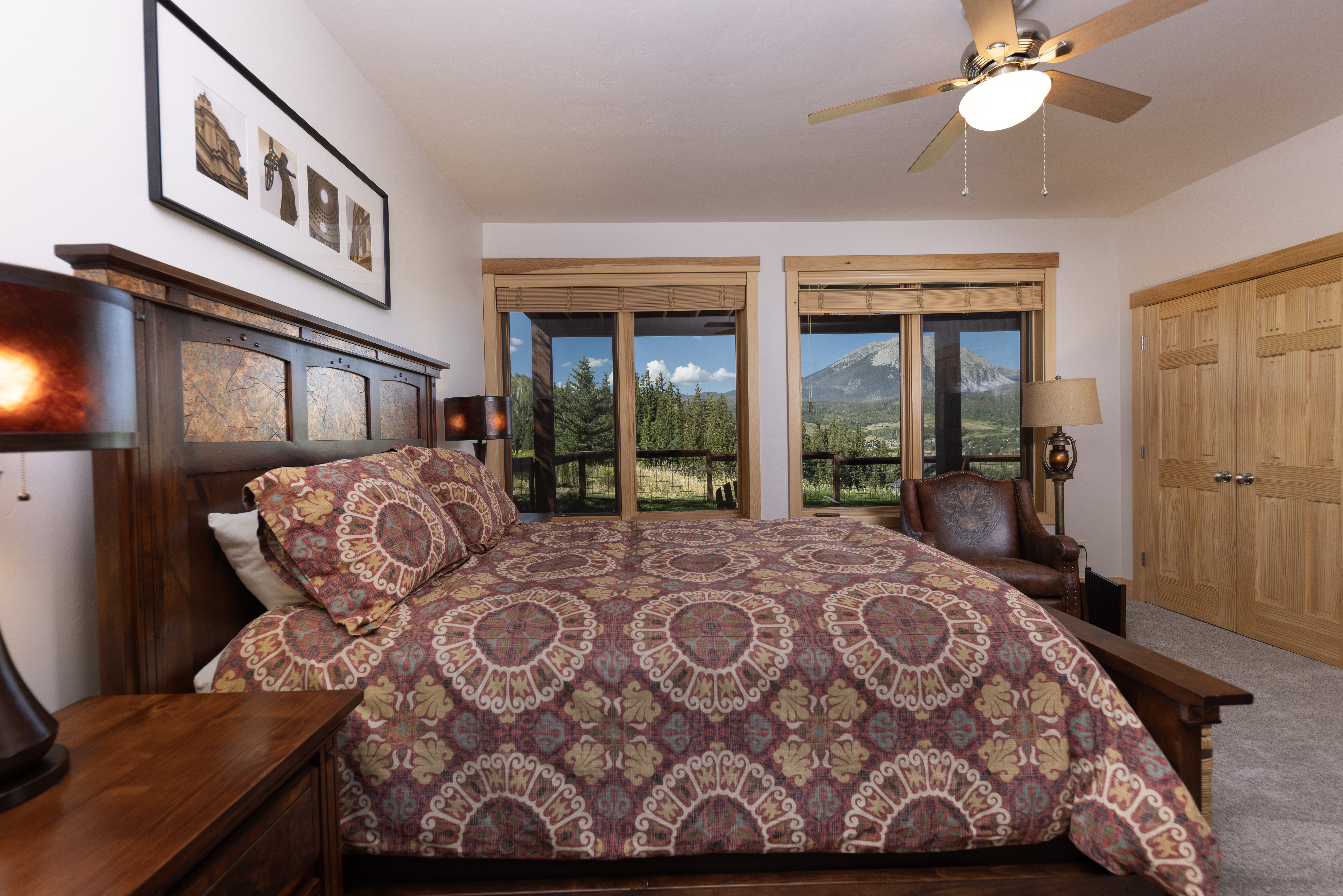This primary bedroom features a rustic aesthetic with a heavy wooden bed frame and a patterned quilt that complements the warm, earthy tones of the room. Large windows frame a scenic mountain view, allowing natural light to fill the space, while a leather armchair sits in the corner for a cozy reading nook. The room is completed by a ceiling fan, matching bedside lamps, and a framed art piece above the headboard, creating a comfortable and inviting atmosphere.