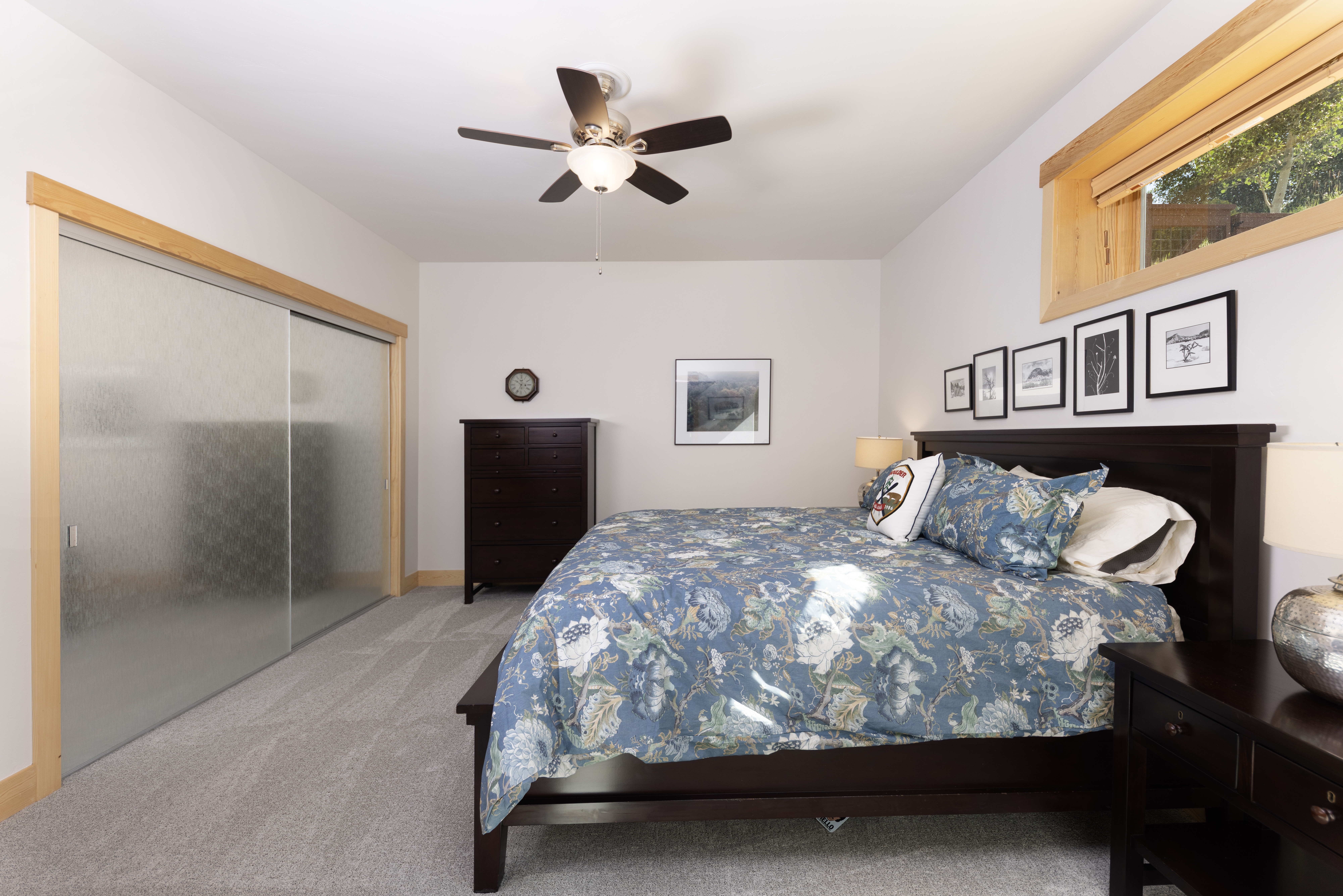 This primary bedroom features a dark wood bed frame dressed in a blue floral patterned comforter and a matching headboard. The room includes a tall dark wood dresser, a matching nightstand with a lamp, and a large sliding-door closet with frosted glass panels. A ceiling fan provides overhead lighting, and a series of framed art pieces hangs above the bed, complemented by a window with light wood trim.