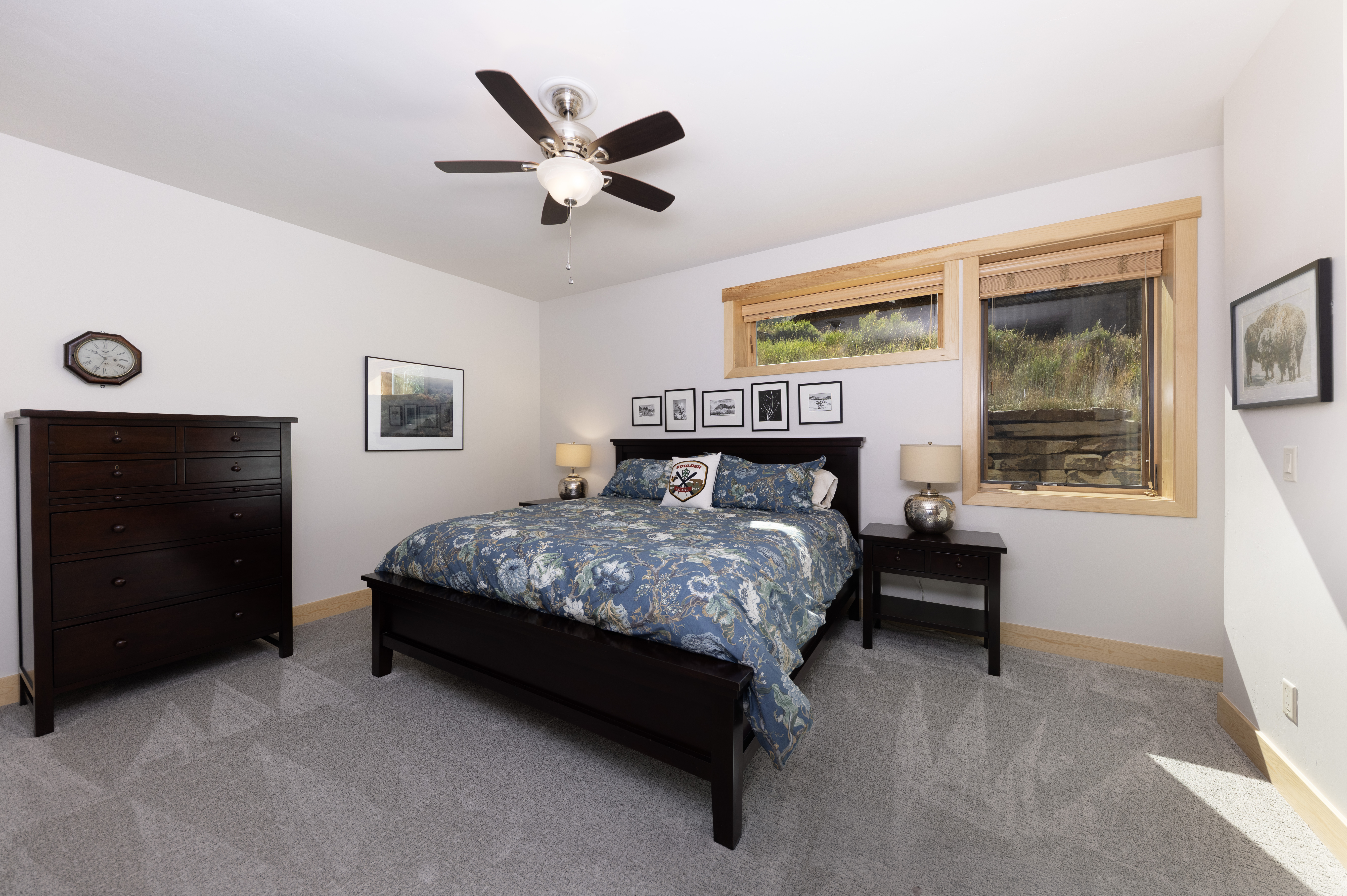This well-lit primary bedroom features a dark wood bed frame with a floral-patterned duvet and a collection of framed wall art above the headboard. The room includes a matching dark wood dresser, two nightstands with lamps, and large windows that provide a view of the natural landscape outside. The space is finished with neutral walls and light-colored carpeting, creating a calm and inviting atmosphere.