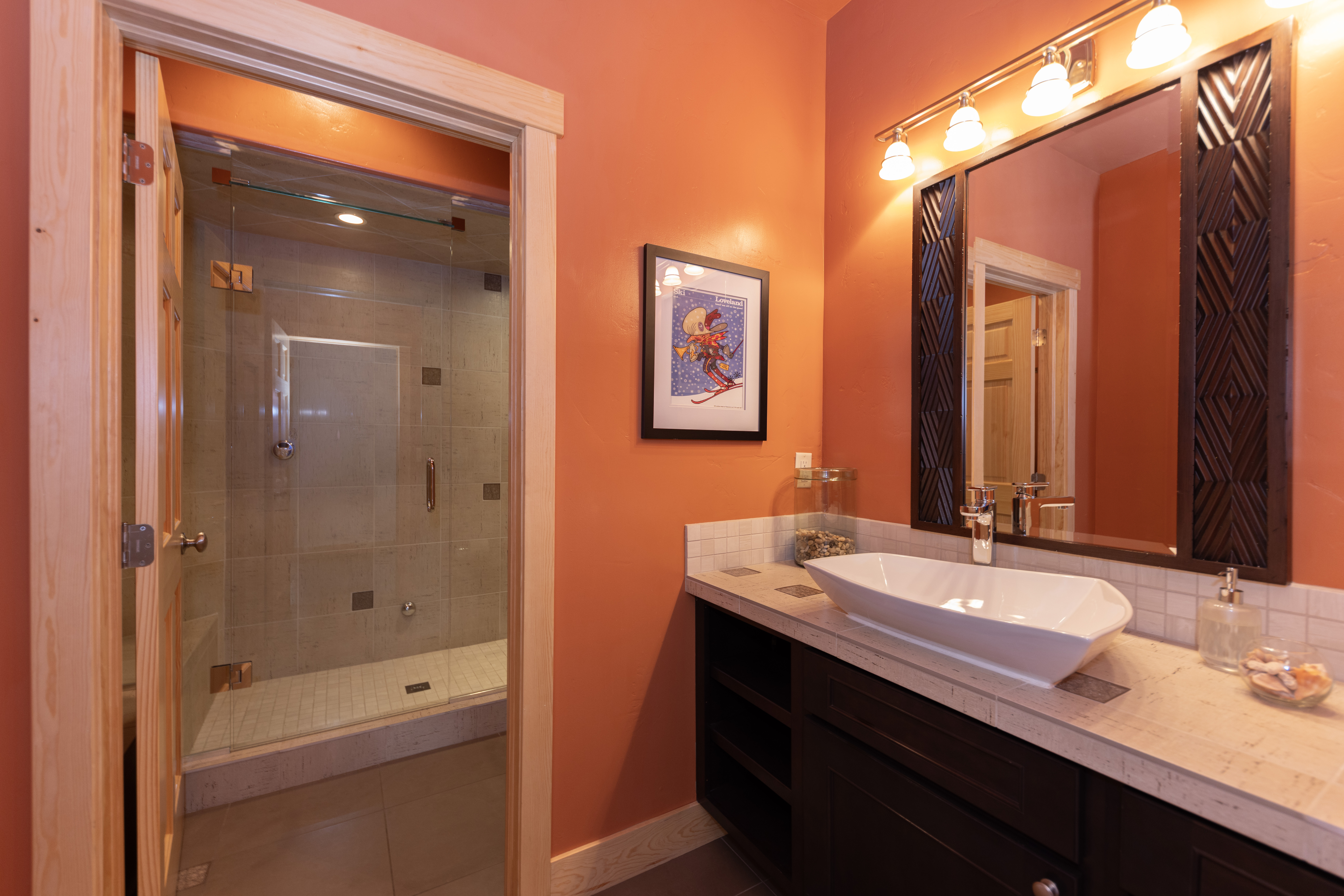 This vibrant bathroom features bold orange walls and a dark wood vanity topped with a light-toned stone counter and a modern vessel sink. A glass-enclosed walk-in shower with light-colored tile is visible through the doorway, while a decorative framed print adds a touch of personality to the space. The room is illuminated by a multi-bulb vanity light fixture mounted above a mirror with dark, textured side panels.