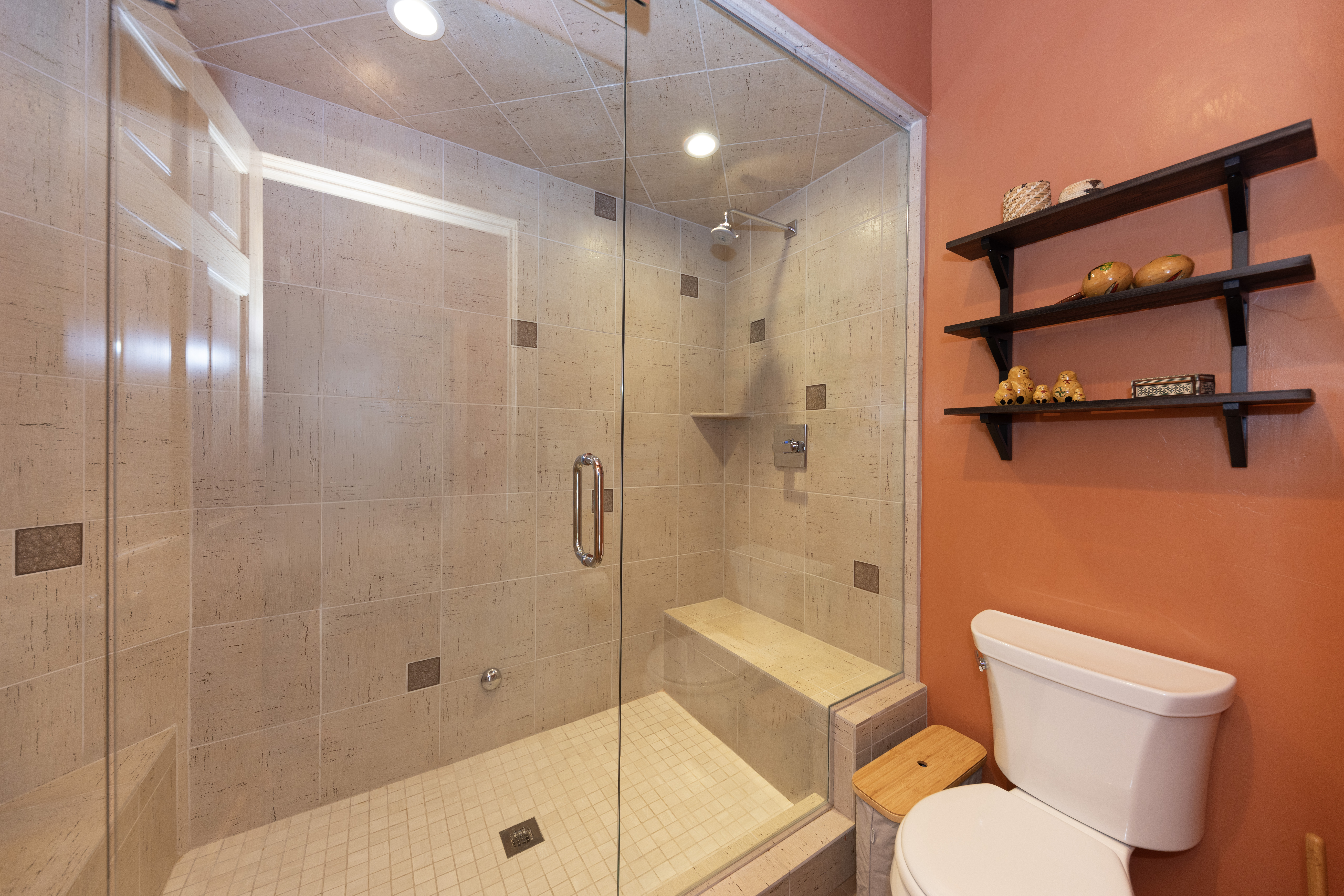 This bathroom features a walk-in shower enclosed by glass panels, showcasing light-toned, textured wall tiles and a built-in bench. The adjacent wall is painted a vibrant orange, providing a bold contrast to the neutral shower area, and is accented by a three-tiered dark wood wall shelf. A white toilet is positioned in the foreground, completing this compact and functional bathroom space.