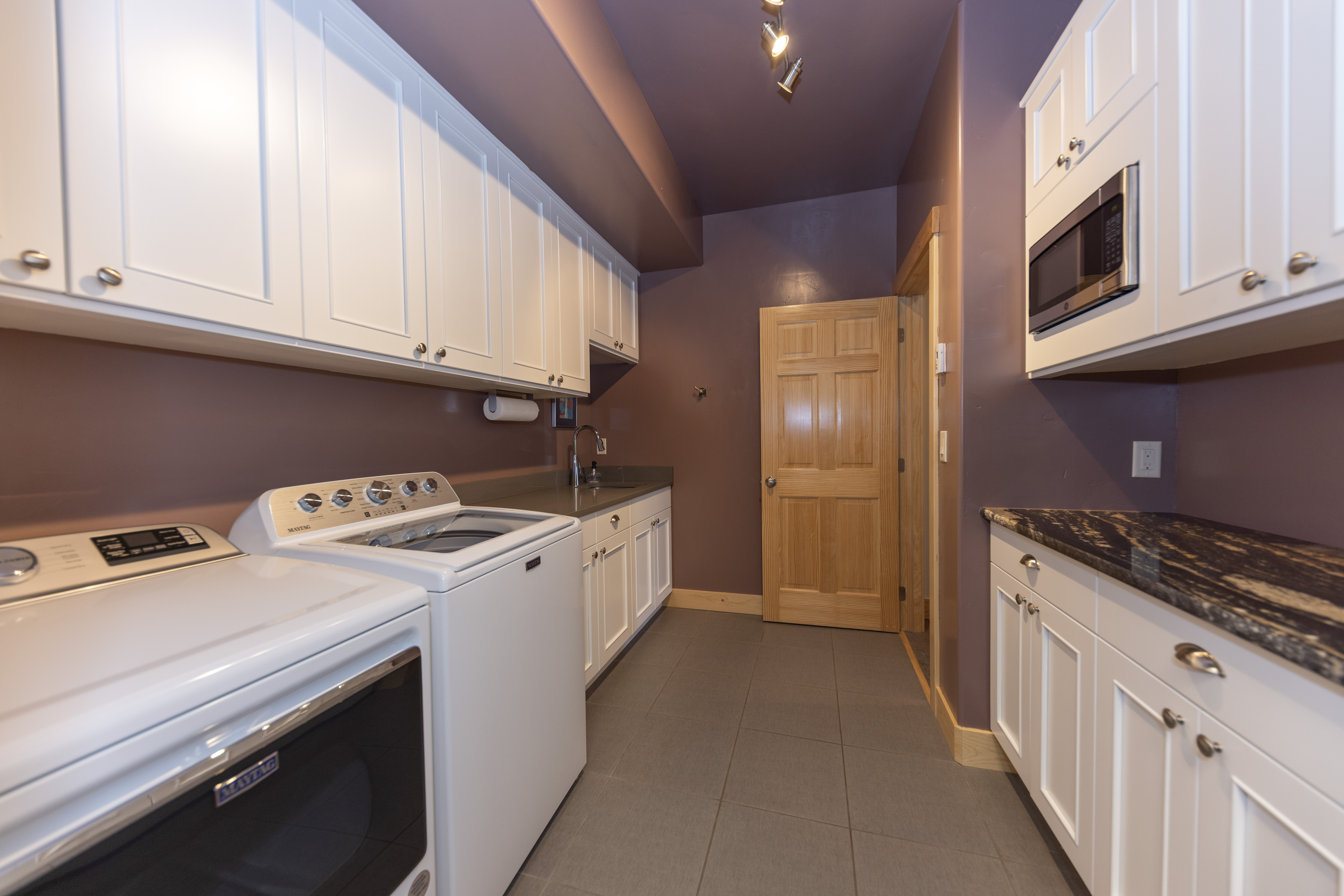 This functional laundry room features white cabinetry, a Maytag washer and dryer set, and a small sink area with a dark countertop. The room is painted in a deep purple hue, contrasted by light wood cabinetry and a natural wood door, creating a distinct and practical workspace. The perspective is a wide-angle shot from the entrance, showcasing the efficient layout and storage capacity of the room.