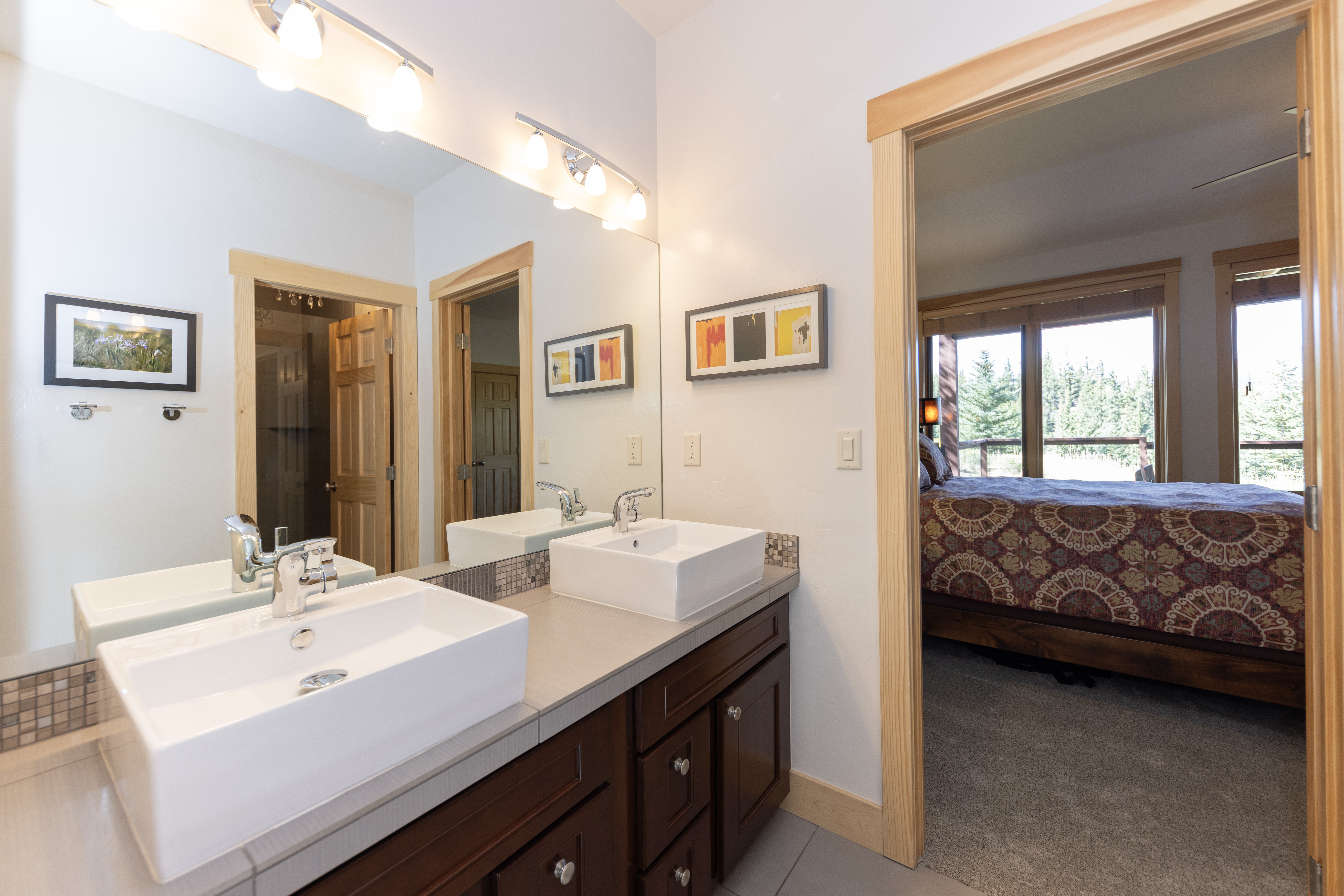 This primary bathroom features a dual-sink vanity with rectangular vessel sinks set atop a neutral stone countertop and dark wood cabinetry. A large, wall-to-wall mirror reflects the space, including a view into an adjacent bedroom with a patterned bedspread and large windows overlooking a wooded area. The room is bright and functional, with light-colored walls and wood trim framing the doorways.