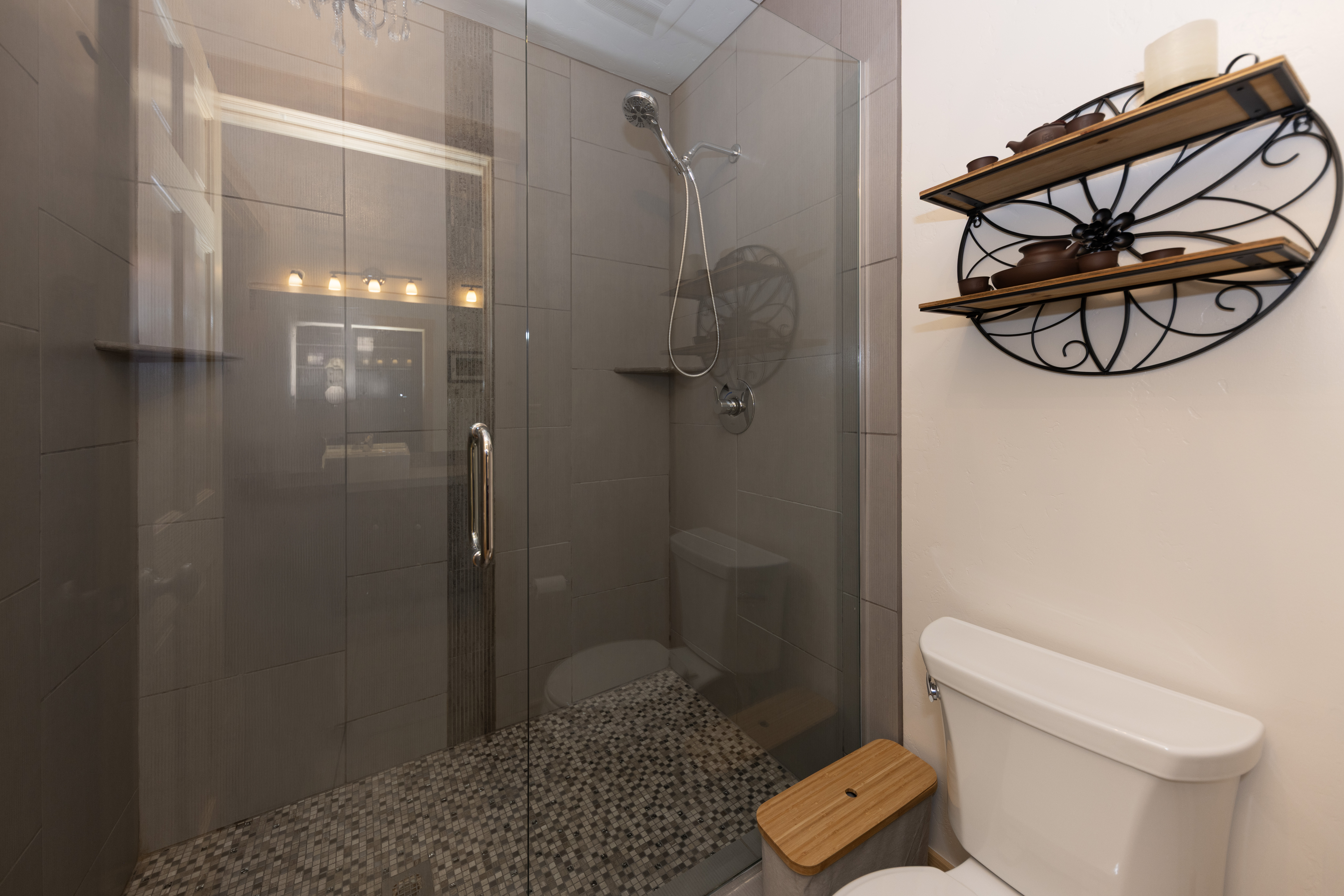 This bathroom features a glass-enclosed walk-in shower with neutral-toned wall tiles and a mosaic-tiled floor. To the right, a white toilet is positioned next to a decorative wall-mounted shelf unit with a circular metal frame and wood shelving. The perspective is a straight-on view capturing the functional layout of the compact space.