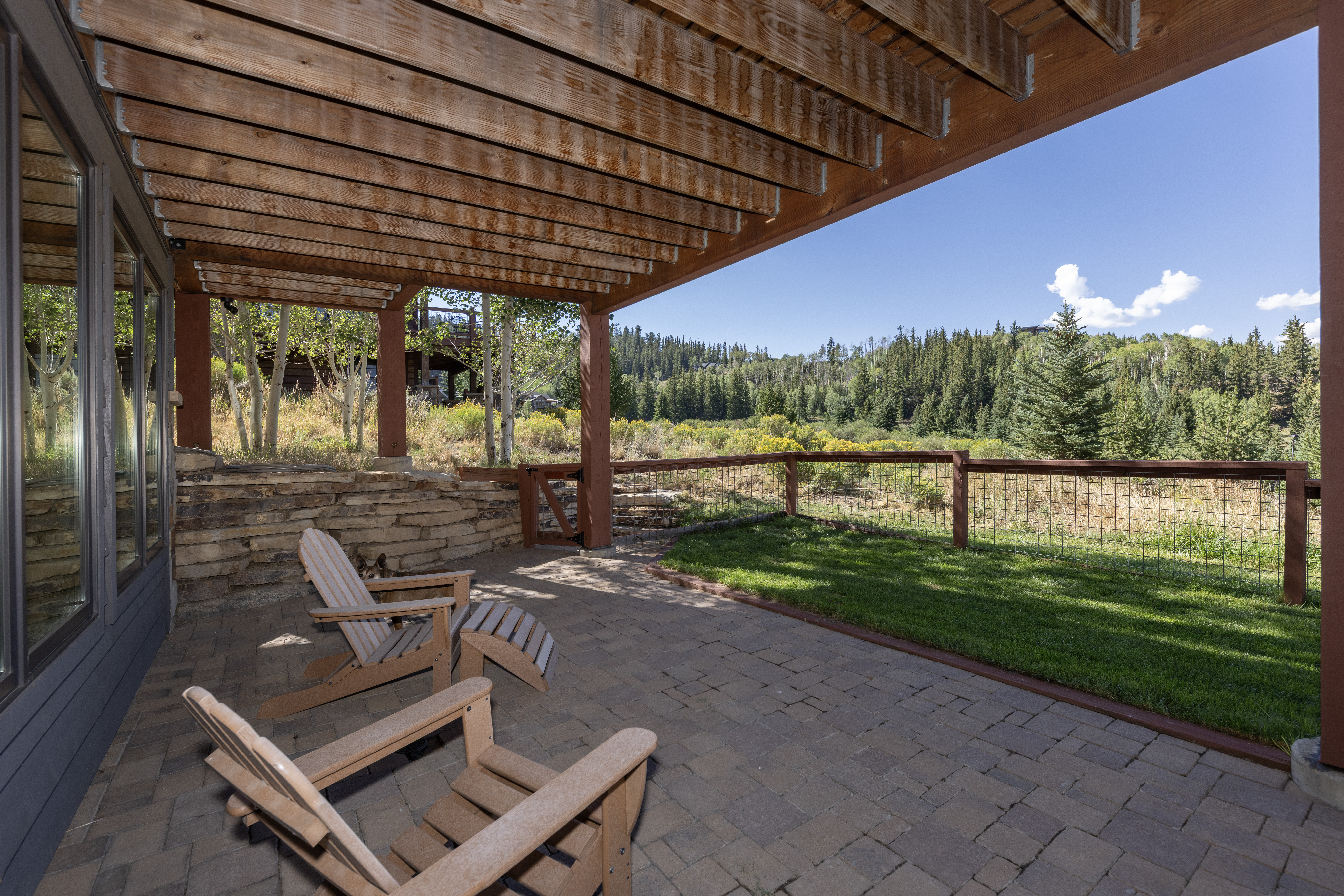 This covered stone patio offers a serene outdoor living space with two Adirondack-style chairs and a matching footrest. The area is framed by a rustic timber pergola and a low stone retaining wall, leading out to a small patch of manicured lawn and a wire-mesh fence that overlooks a scenic, forested mountain landscape. The perspective captures the transition from the shaded, comfortable seating area to the expansive, sun-drenched natural surroundings.