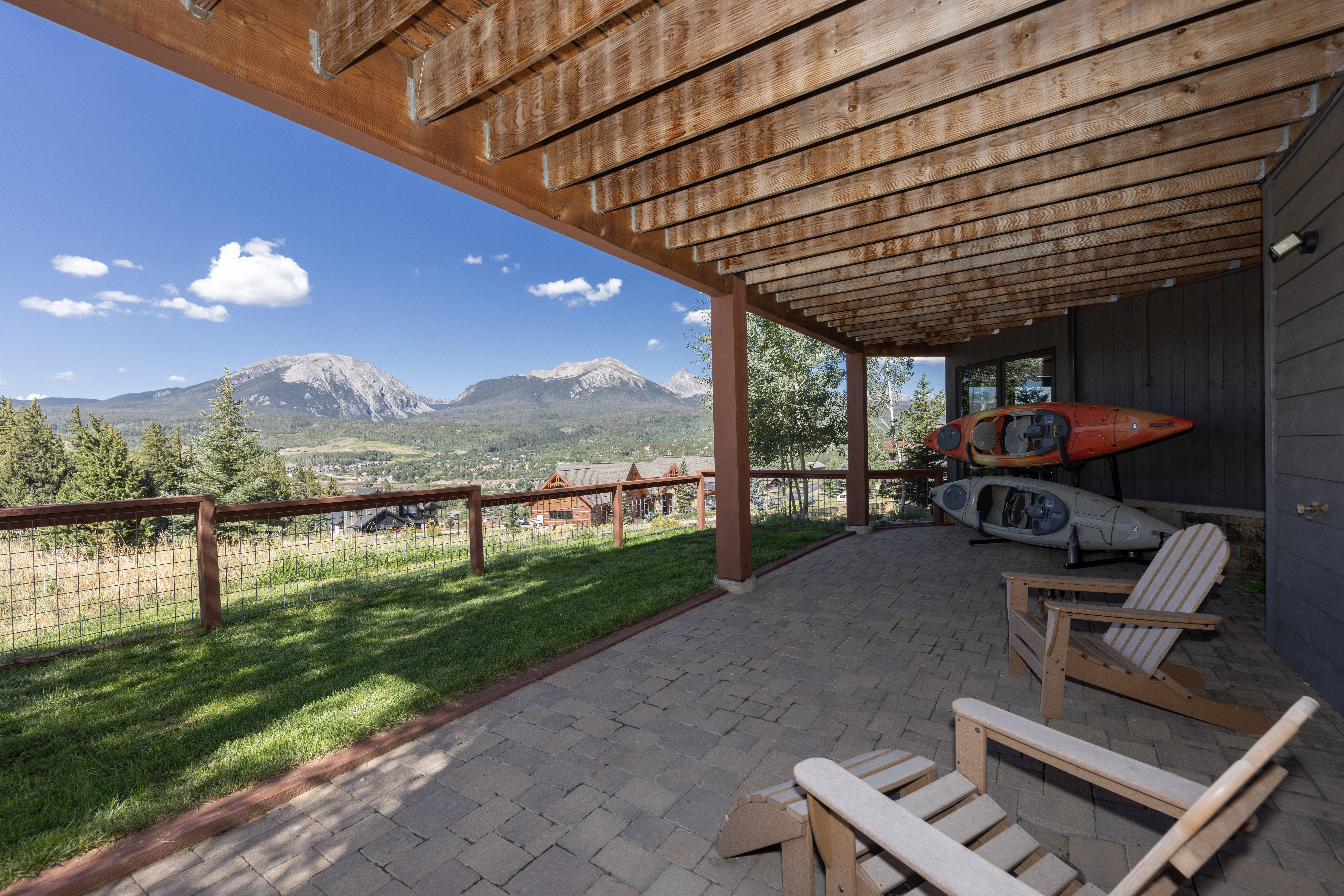 This covered patio features a rustic wood-beam ceiling and a paved stone floor, offering a comfortable outdoor living space. Two wooden Adirondack chairs are positioned to take in the expansive, panoramic mountain views visible beyond the wire-mesh fence and grassy yard. Two kayaks are neatly stored on a rack against the side of the house, emphasizing the recreational lifestyle of the property.