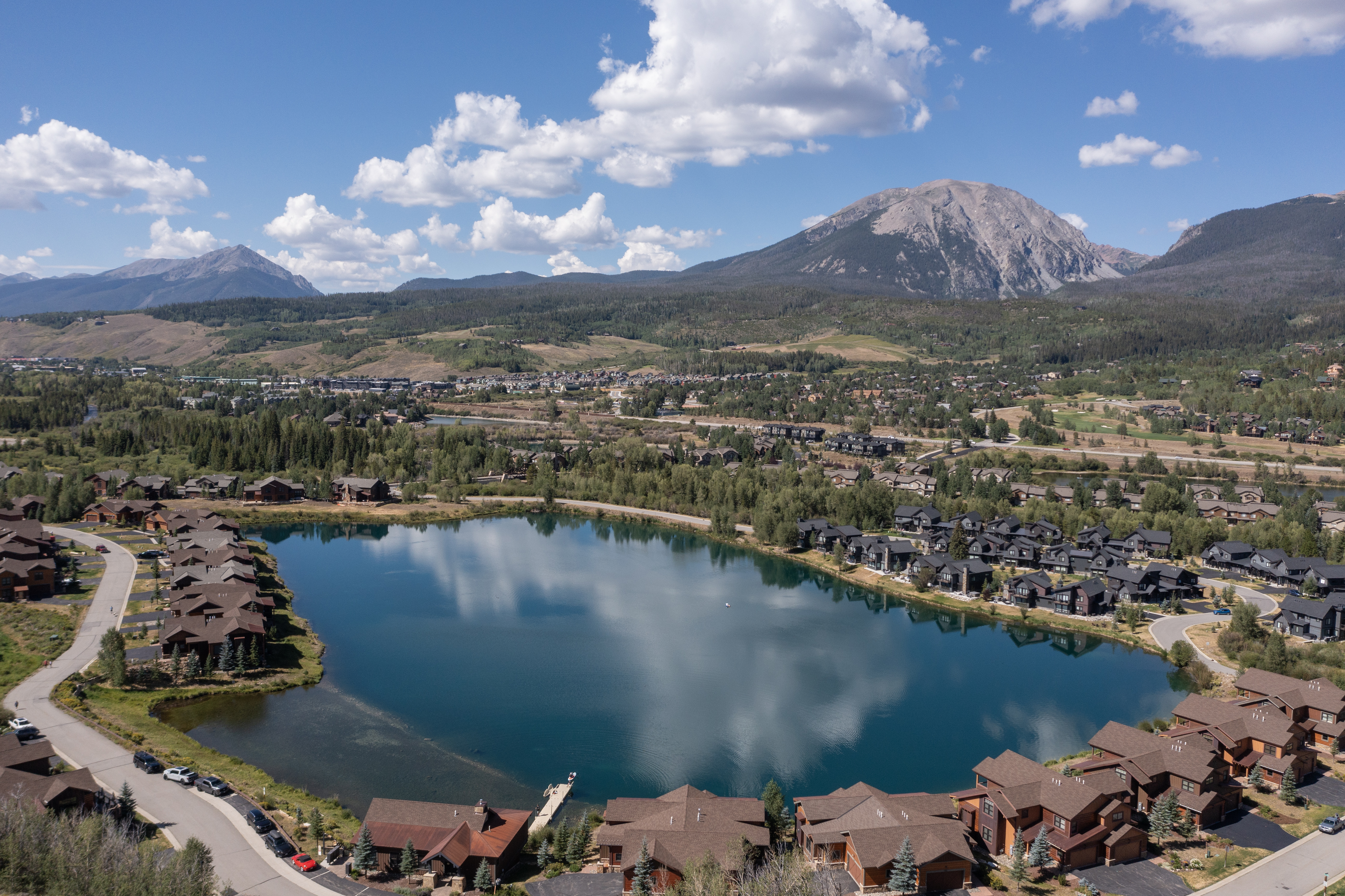 This high-altitude aerial view captures a serene residential community nestled around a central, deep blue lake. The landscape features a mix of mountain-style homes with dark roofs and wood-toned siding, surrounded by lush green trees and winding roads. In the background, majestic, rugged mountain peaks rise against a bright blue sky filled with scattered white clouds, creating a picturesque and tranquil setting.