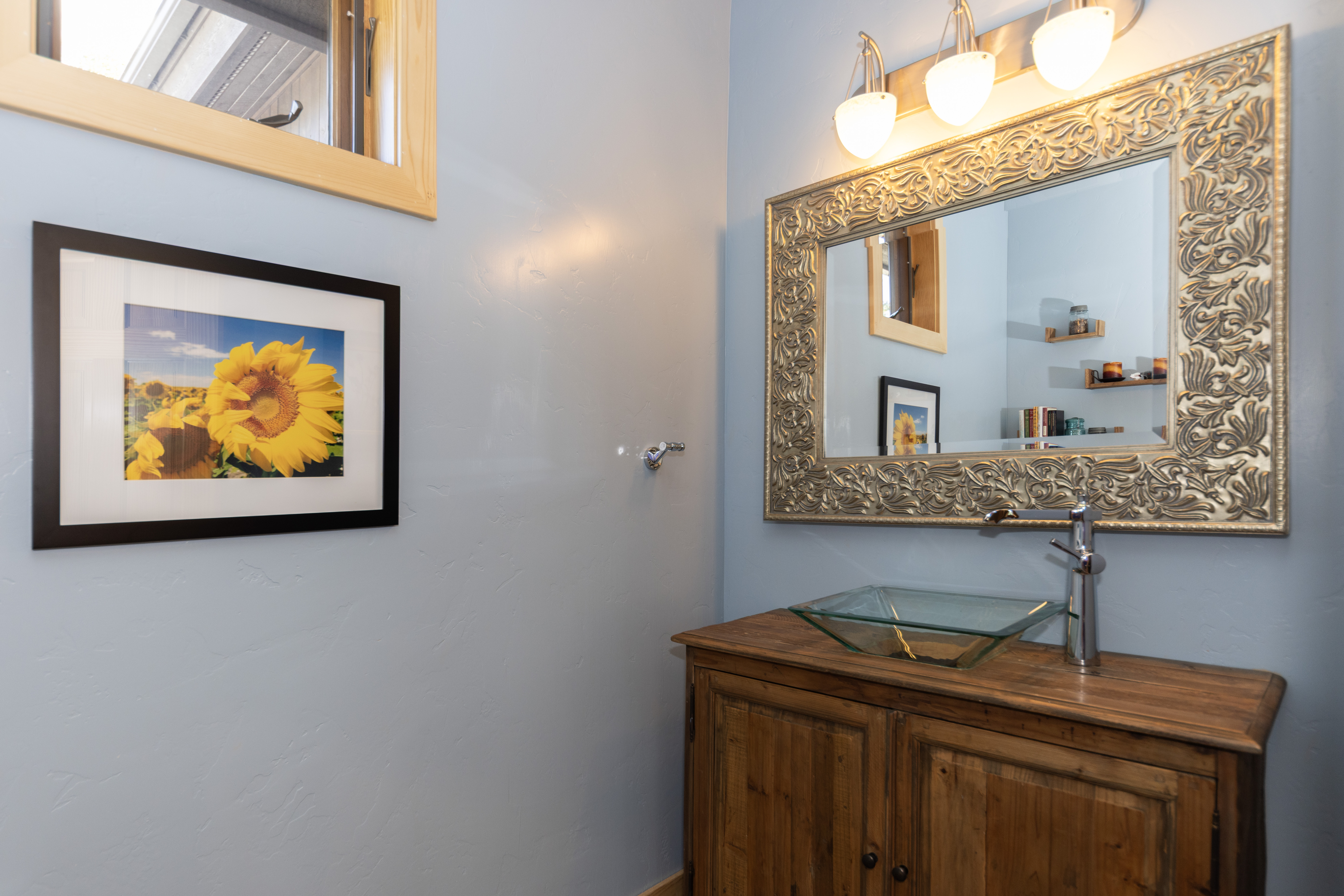This bathroom features a rustic wooden vanity topped with a unique glass vessel sink and a modern chrome faucet. A large, ornate metallic-framed mirror hangs above the vanity, reflecting the light from a three-bulb fixture and a framed sunflower print on the adjacent light blue wall. The space is accented by a small window with a light wood frame, creating a warm and inviting atmosphere.