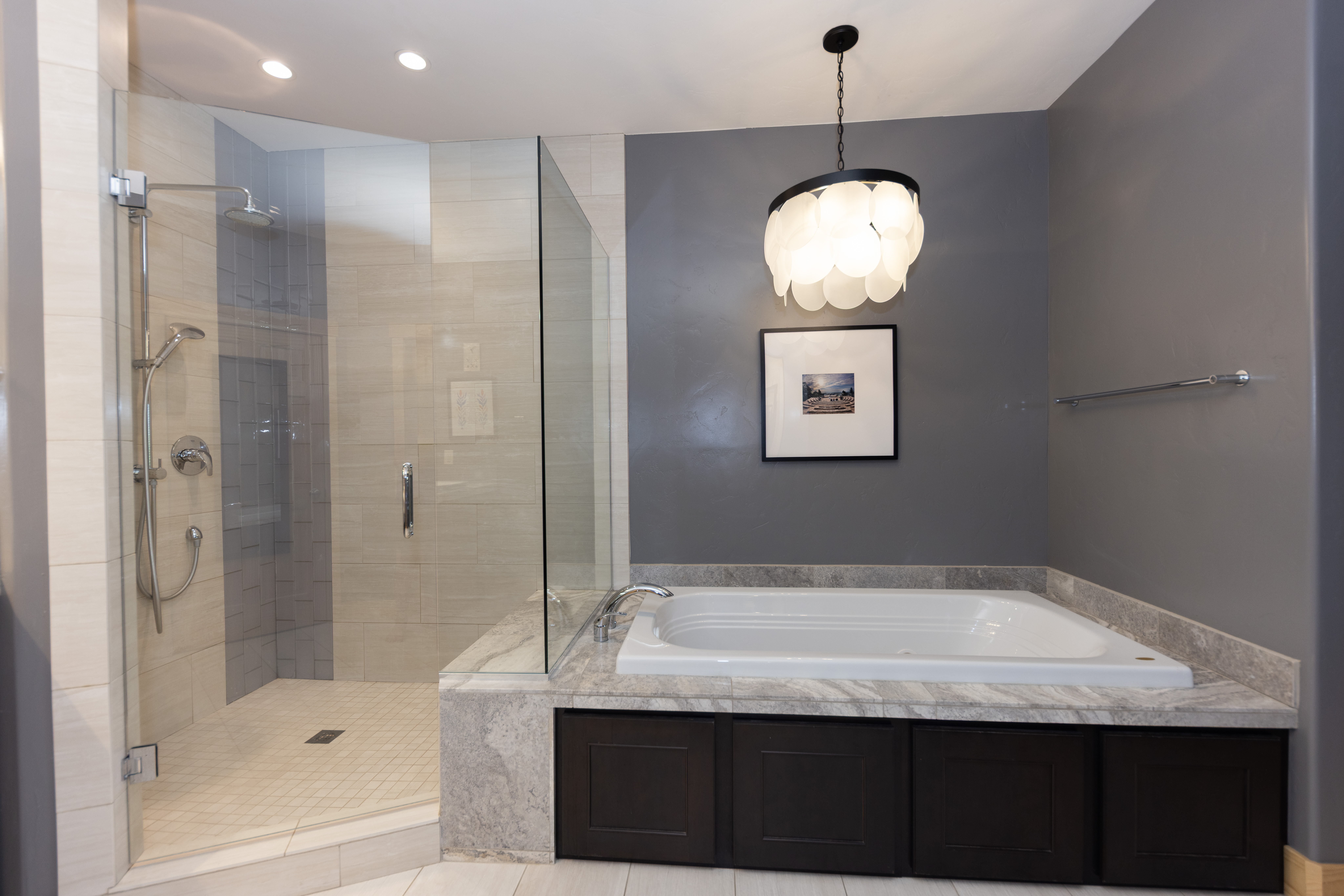 This primary bathroom features a spacious walk-in shower with glass panels and a separate soaking tub set into a stone-topped vanity with dark cabinetry. A decorative, multi-layered pendant light hangs above the tub, complemented by a framed piece of art on the dark gray accent wall. The space combines neutral tile flooring and wall accents with a sophisticated, high-contrast color palette.