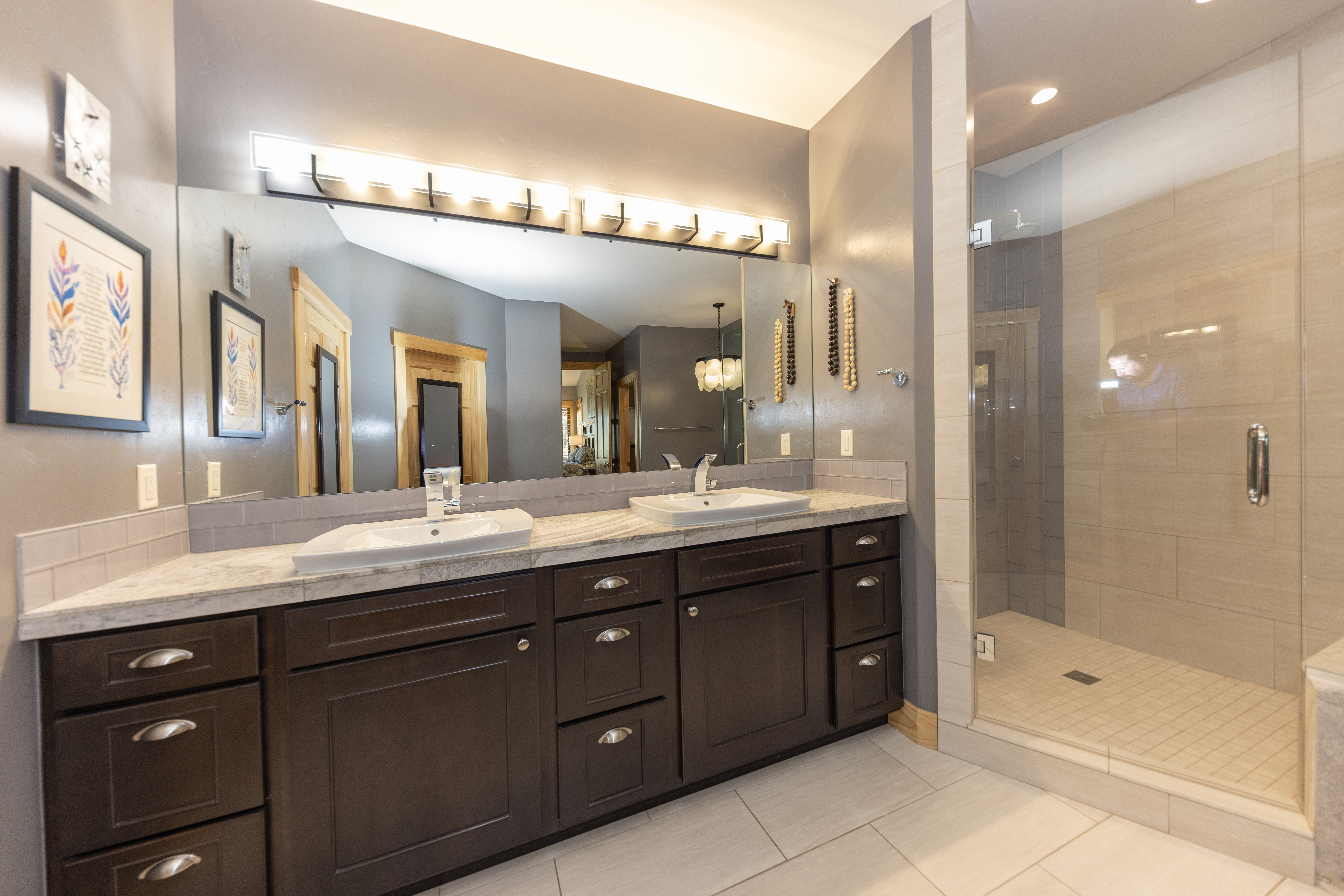 This primary bathroom features a double vanity with dark wood cabinetry, stone countertops, and two rectangular vessel sinks. A large, wide mirror spans the wall above the vanity, illuminated by a multi-bulb light fixture, while a glass-enclosed walk-in shower with neutral-toned wall tile sits to the right. The space is finished with light-colored floor tiles and gray walls, creating a clean and functional aesthetic.