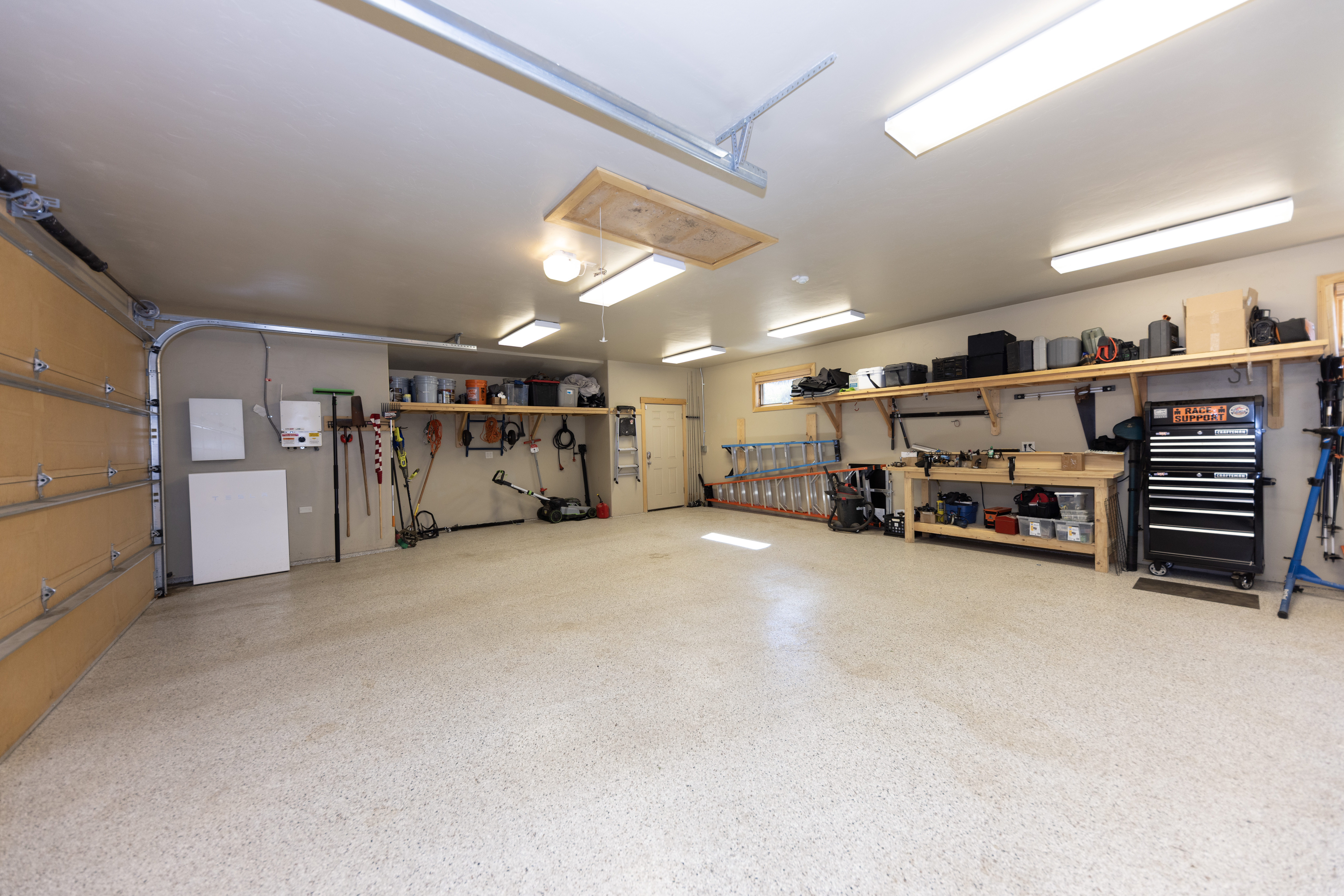 This spacious, well-organized garage features a durable speckled epoxy floor and ample storage solutions, including wall-mounted shelving and a dedicated workbench. The area is brightly lit by multiple overhead fluorescent fixtures, highlighting the clean, functional layout suitable for both vehicle storage and workshop activities. A side door provides convenient access, while the open space offers versatility for various projects or storage needs.