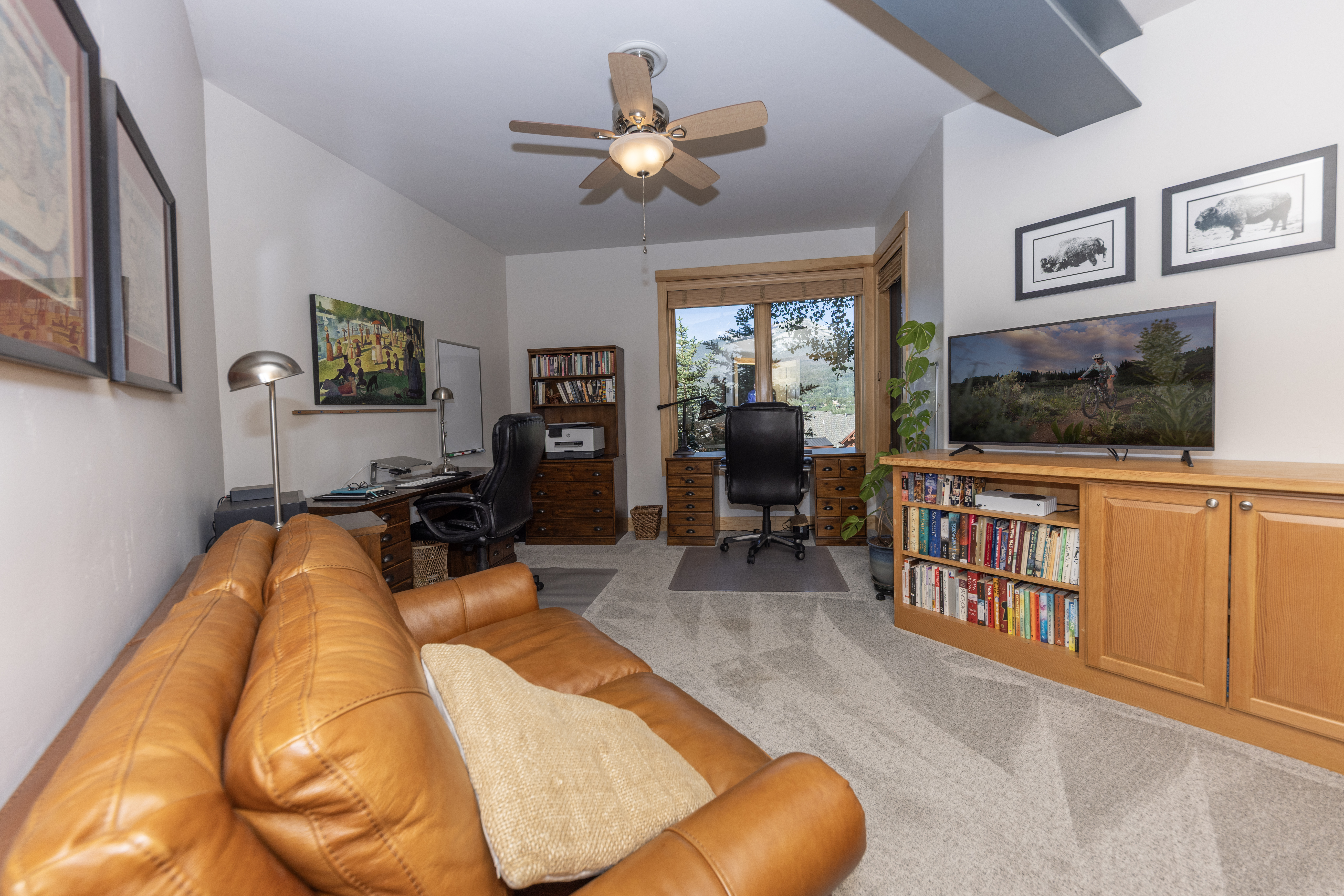 This functional home office features a warm, inviting atmosphere with a leather sofa in the foreground and two distinct desk areas. The room is equipped with built-in wooden cabinetry, bookshelves, and a television, while large windows provide a scenic view of the outdoors. The space is well-lit by a ceiling fan with integrated lighting and a desk lamp, creating a comfortable environment for work or relaxation.