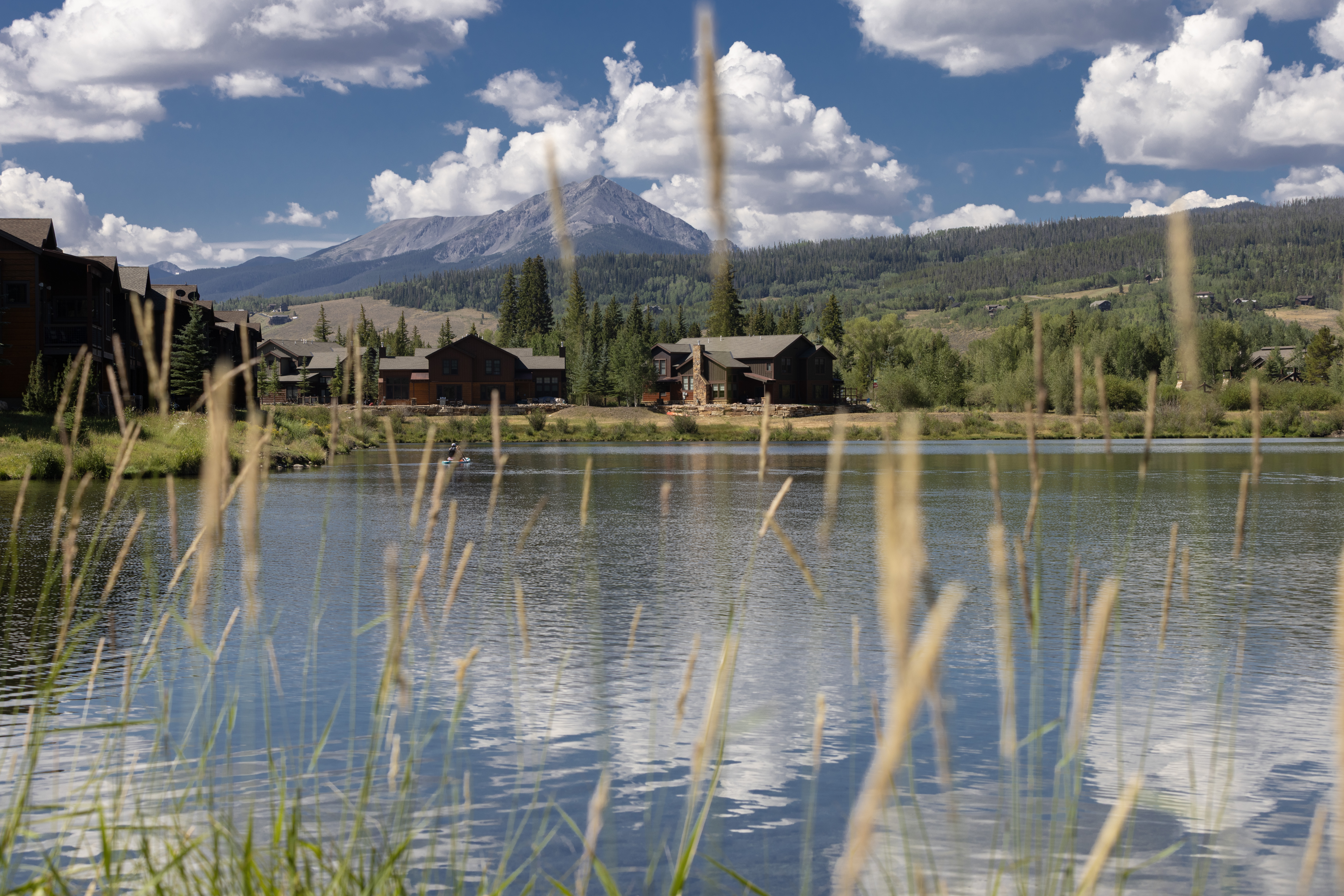 This scenic view captures a tranquil lakefront setting featuring multi-story residential buildings nestled against a backdrop of lush pine forests and a prominent mountain peak. In the foreground, tall blades of grass frame the composition, creating a soft, cinematic depth that draws the eye toward the calm water and the architectural structures. The bright, cloud-filled sky reflects beautifully on the lake's surface, emphasizing the serene and picturesque nature of this community.