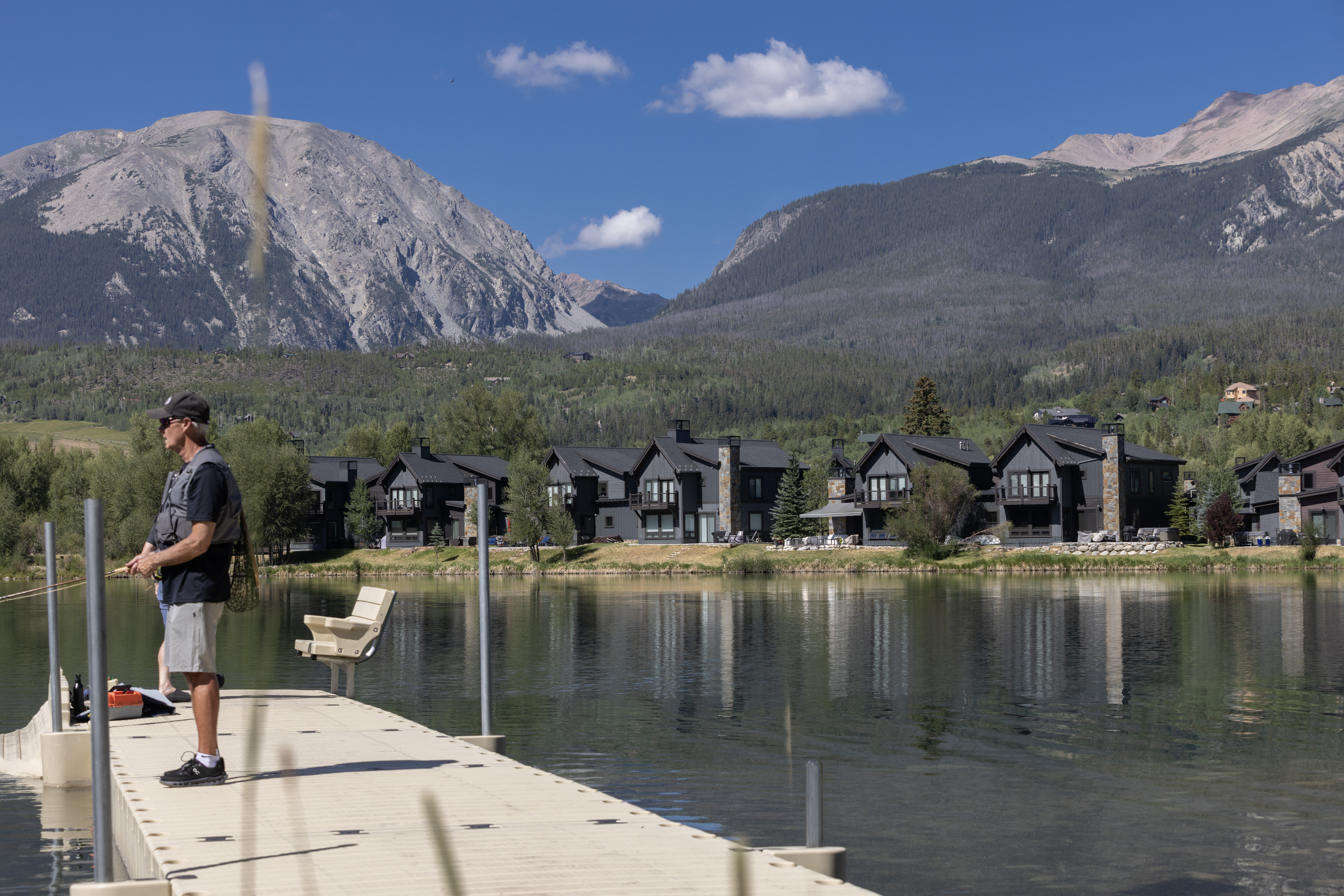 This scenic shot captures a tranquil lakefront setting featuring a series of modern, dark-toned residential homes nestled against a backdrop of majestic, rugged mountains. In the foreground, a person stands on a floating dock, engaged in fly fishing, which emphasizes the recreational lifestyle offered by the community. The composition highlights the peaceful integration of contemporary architecture with the natural landscape, creating an inviting and serene atmosphere.