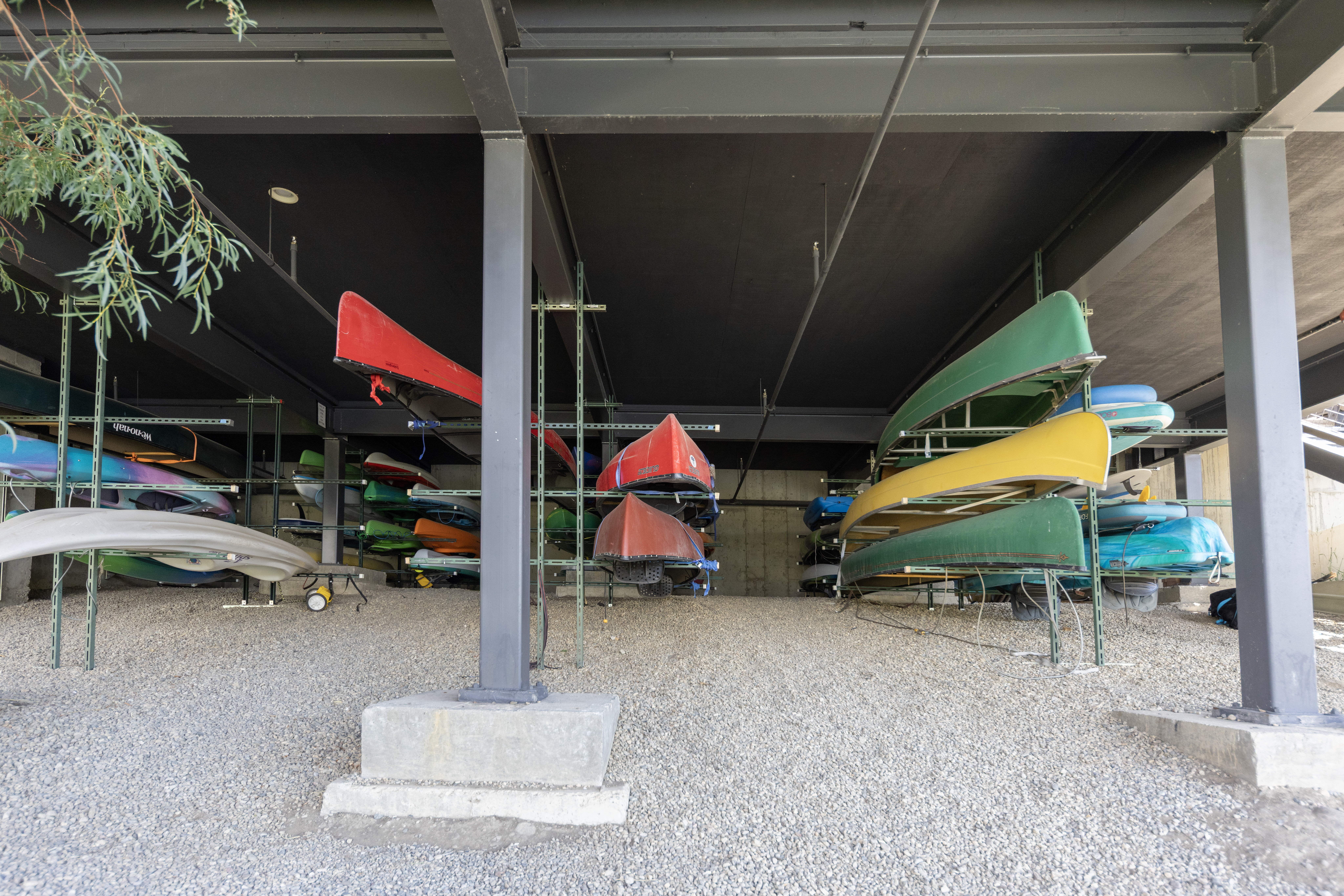 This image captures a covered outdoor storage area designed for watercraft, featuring multiple metal racks holding a variety of colorful canoes and kayaks. The structure is supported by sturdy dark-painted steel beams and columns, set over a gravel ground surface. The perspective is centered, looking into the sheltered space, highlighting the organized storage solution for recreational equipment.