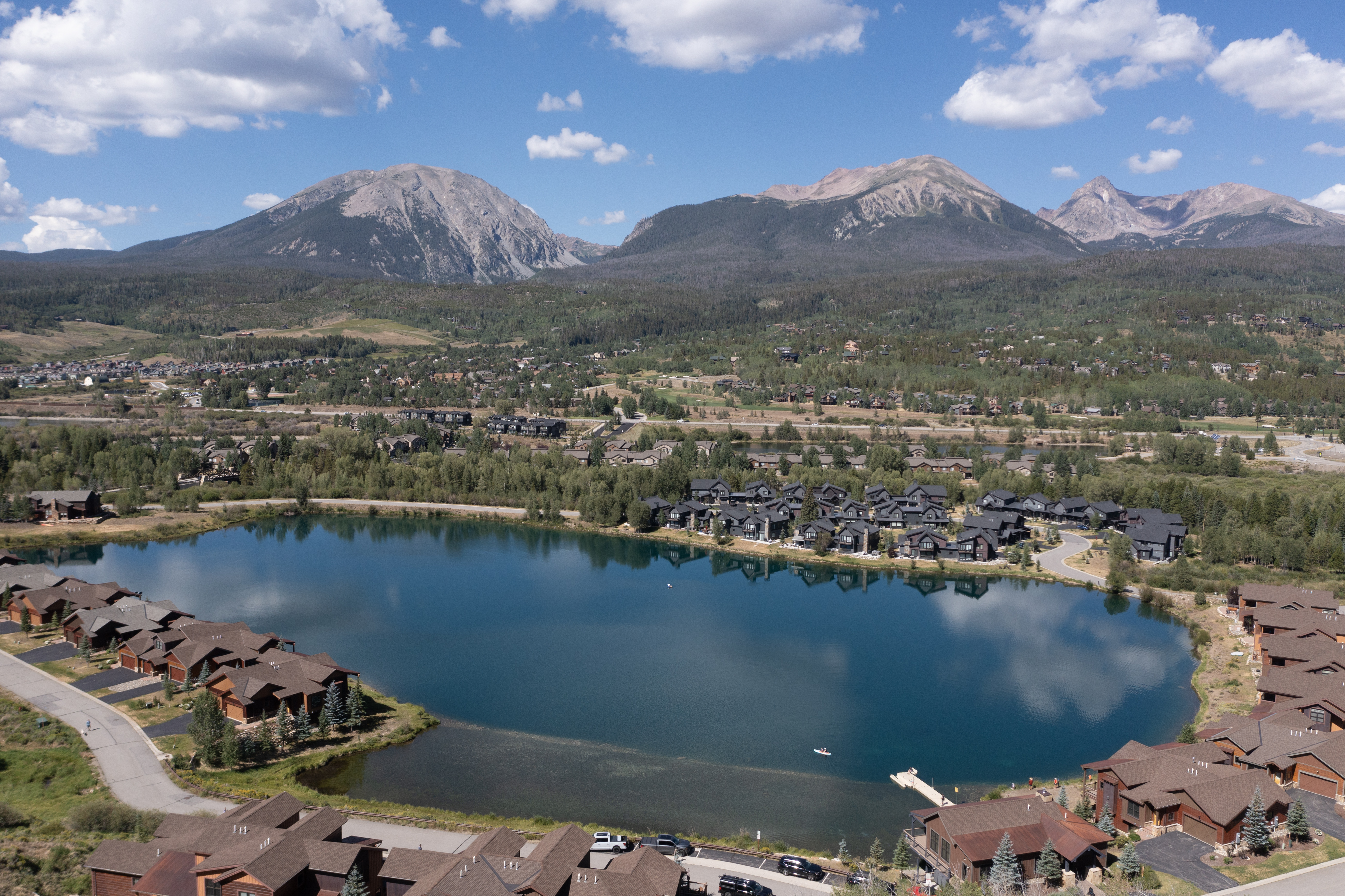 This high-angle aerial view captures a scenic mountain community centered around a tranquil, deep blue lake. The landscape features clusters of multi-story residential homes with dark-toned roofs and wood-accented exteriors, nestled among lush green trees and rolling hills. In the background, majestic, rugged mountain peaks rise against a bright blue sky dotted with soft, white clouds, creating a serene and picturesque setting.